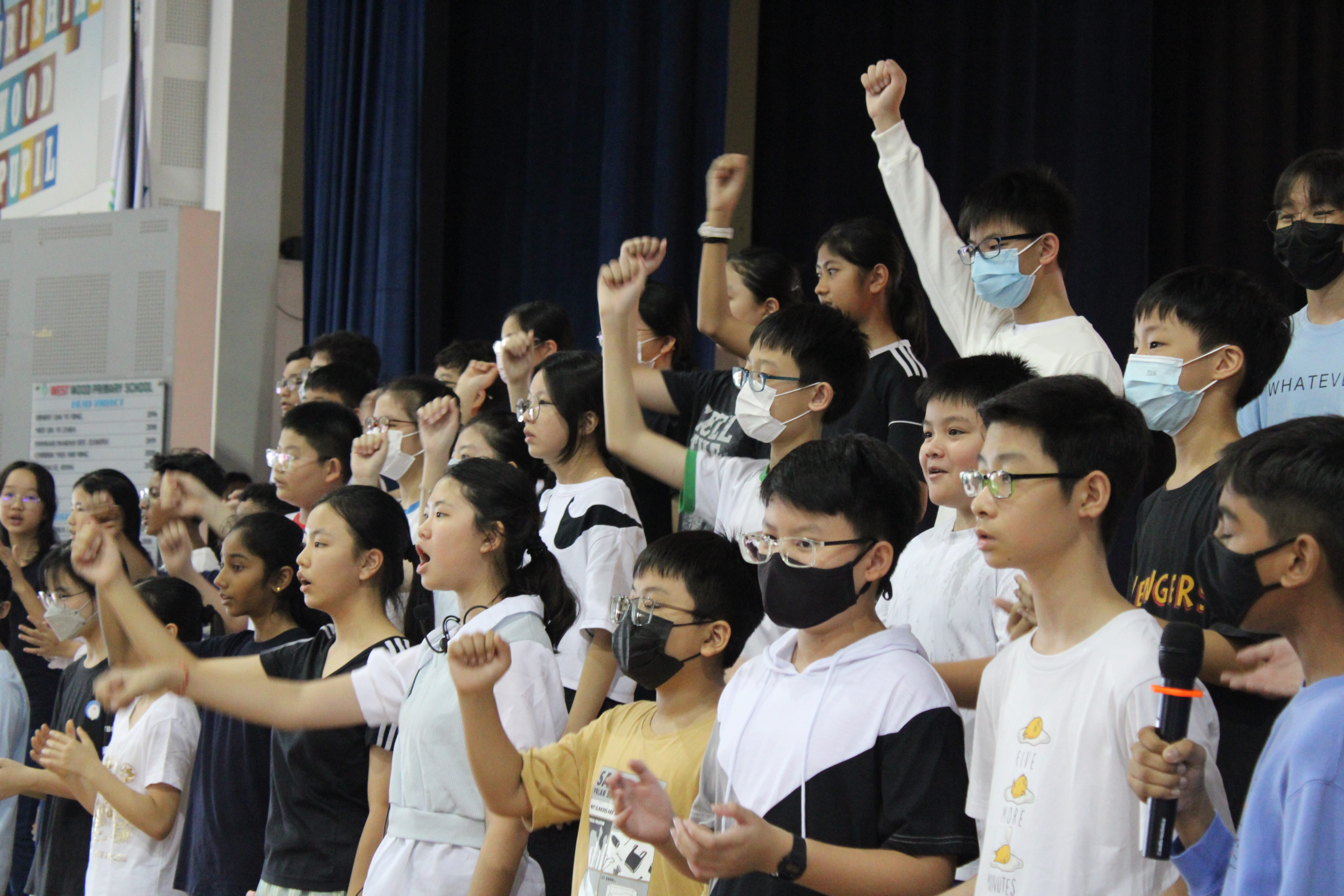 Group of students onstage, many wearing masks, raising fists and singing; one holds a microphone.