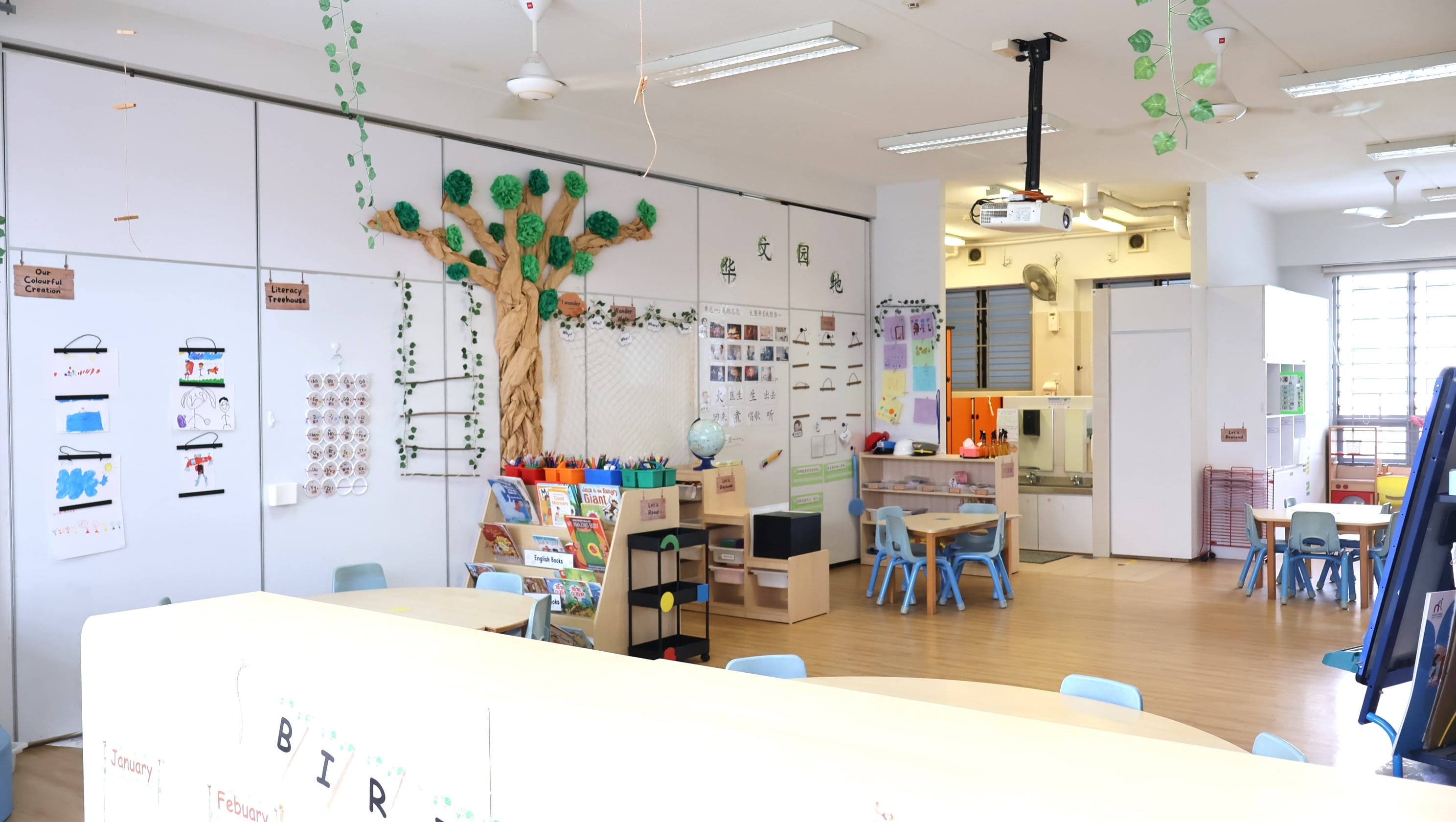 A bright kindergarten classroom with small blue chairs, a tree mural, bookshelves, and Chinese characters on the wall.