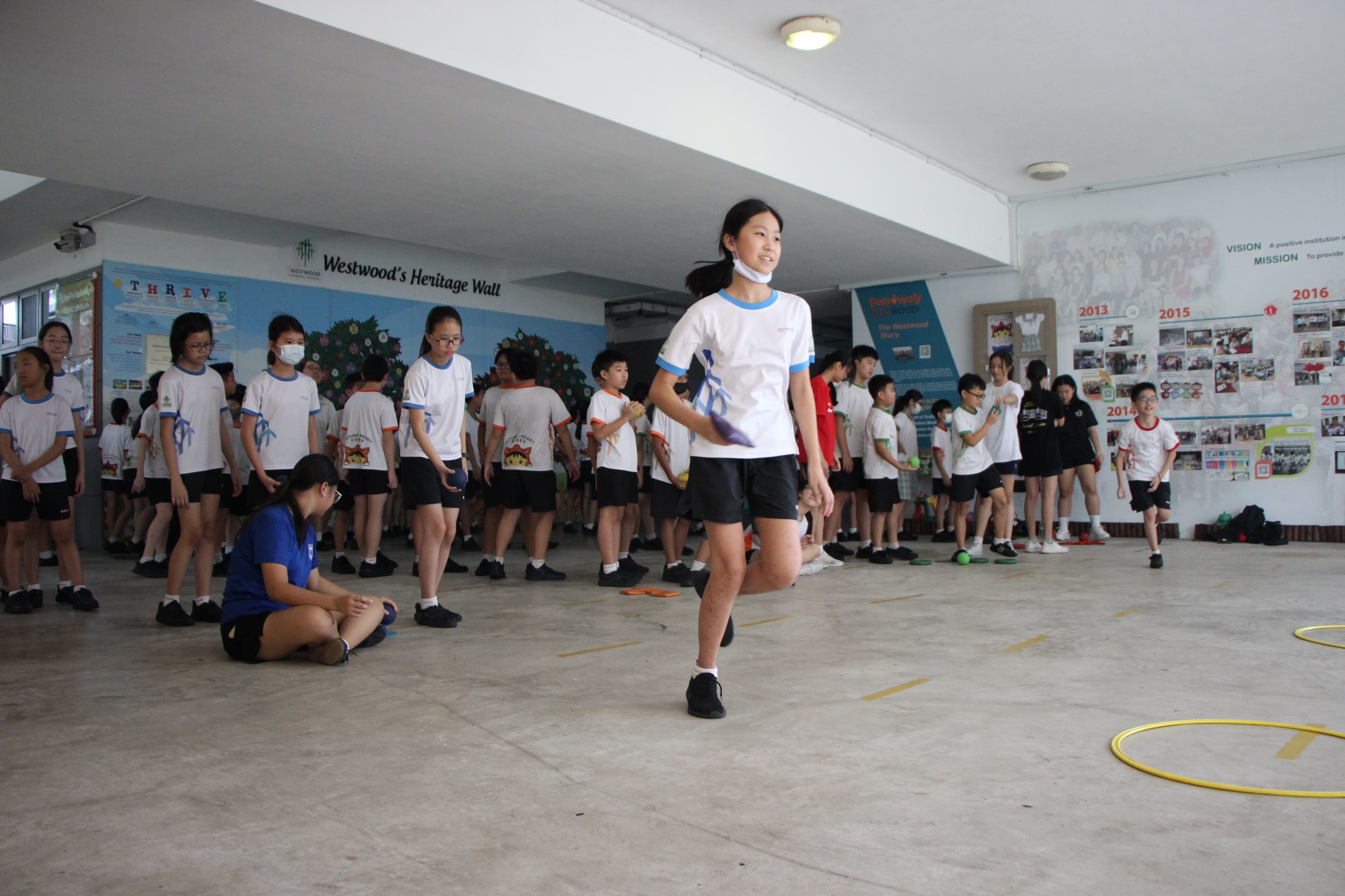 Students in white uniforms participate in an indoor physical activity at Westwood school near a heritage wall display.
