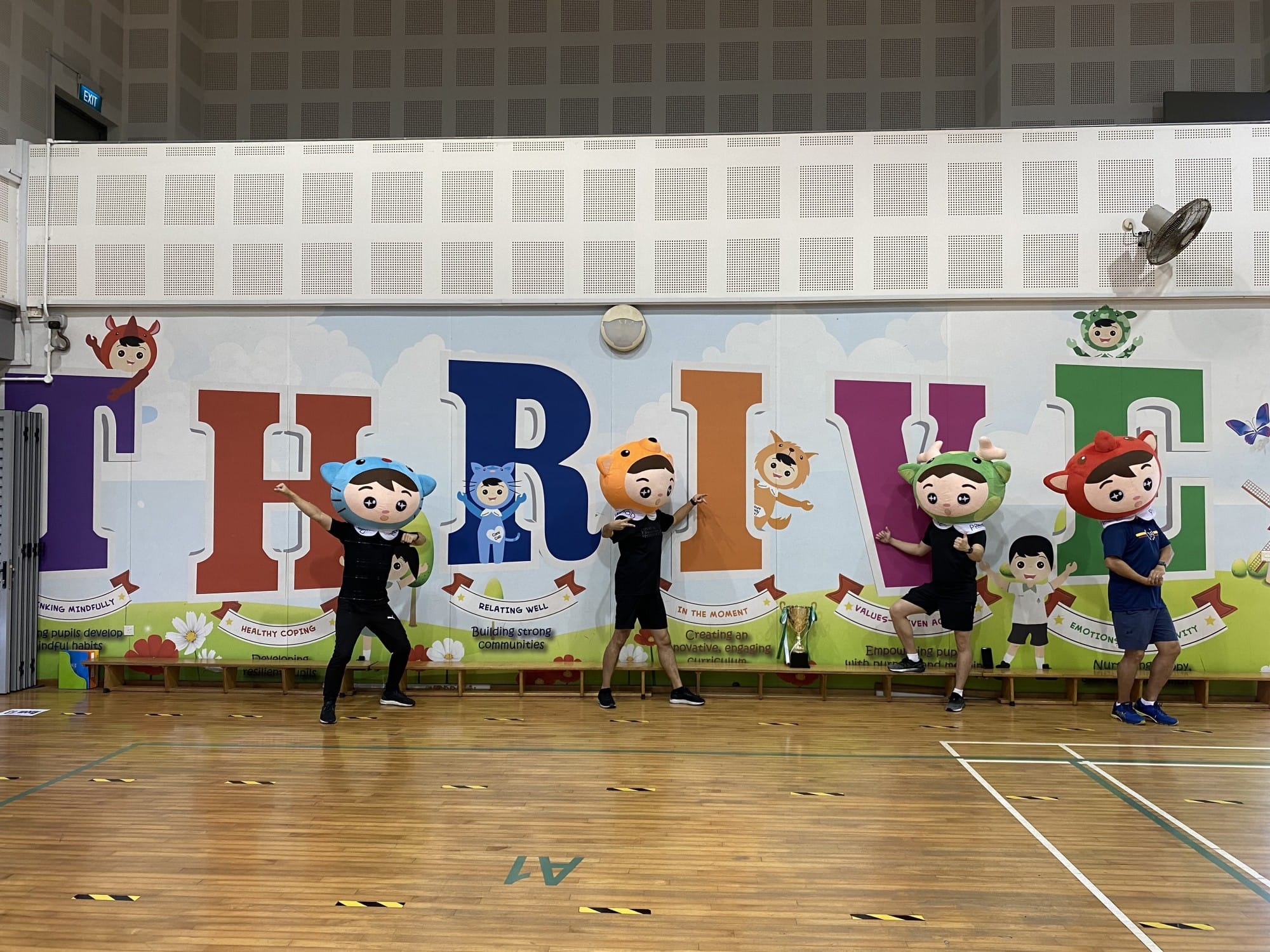 Four performers wearing oversized cartoon mascot heads dance in a gym in front of a colorful THRIVE mural.