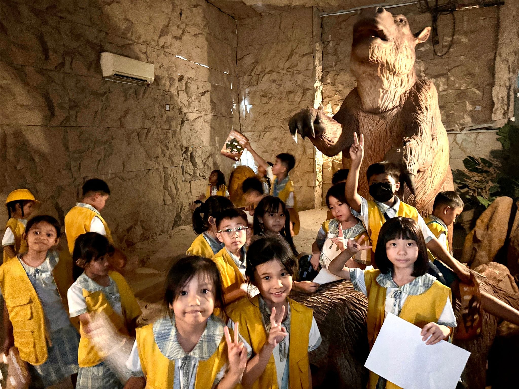 Children in yellow vests pose with peace signs near a large bear statue inside a cave-like museum exhibit.