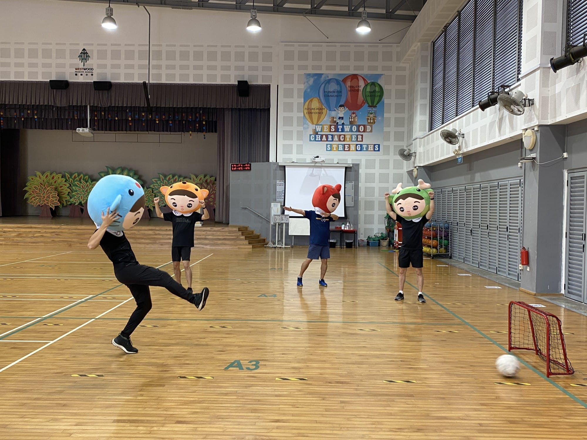 Four people wearing large cartoon character head costumes play indoor soccer in a school gymnasium.