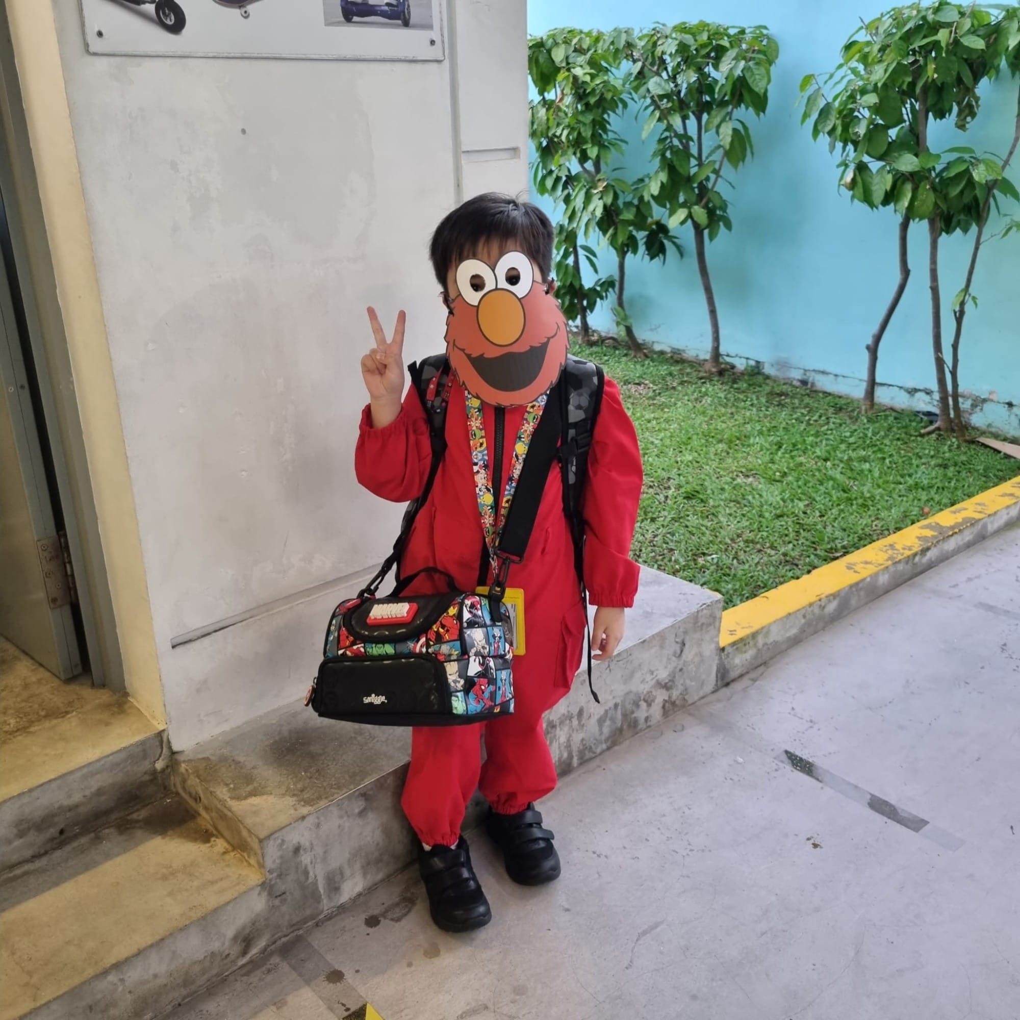 Child in red outfit wearing an Elmo mask, holding a peace sign, with a colorful bag, standing outdoors.