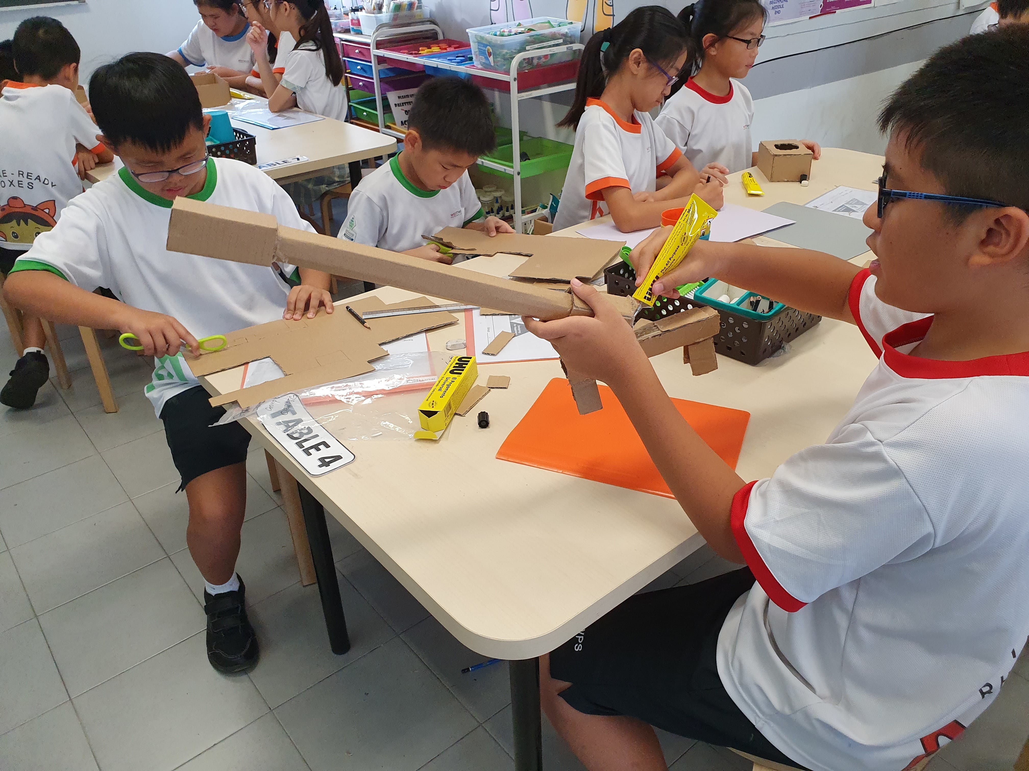 Students in white uniforms build cardboard structures using scissors and glue at classroom tables.