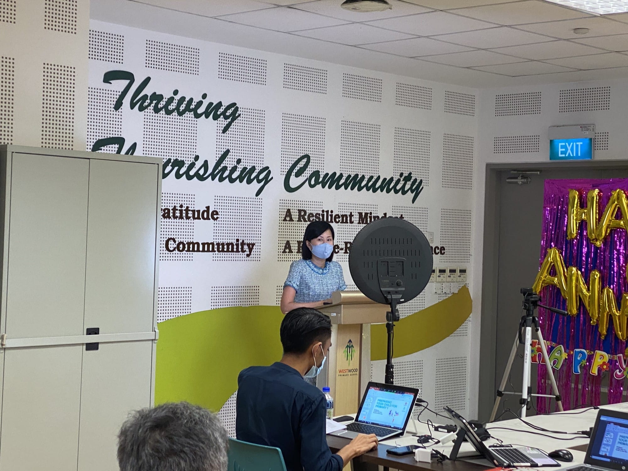 A masked woman speaks at a podium in front of a wall reading "Thriving Community" at Westwood Primary School.