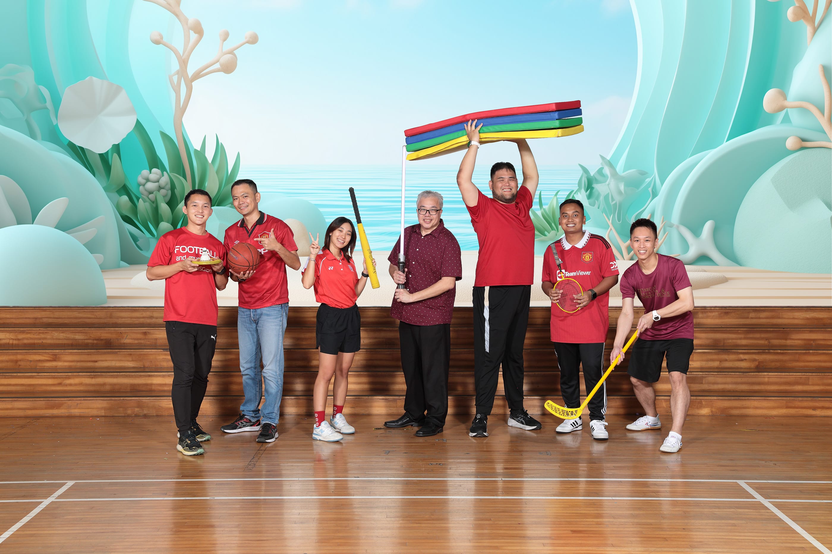 Seven people in red shirts pose on a gym floor holding sports equipment including a basketball, bat, racket, and colorful mats.