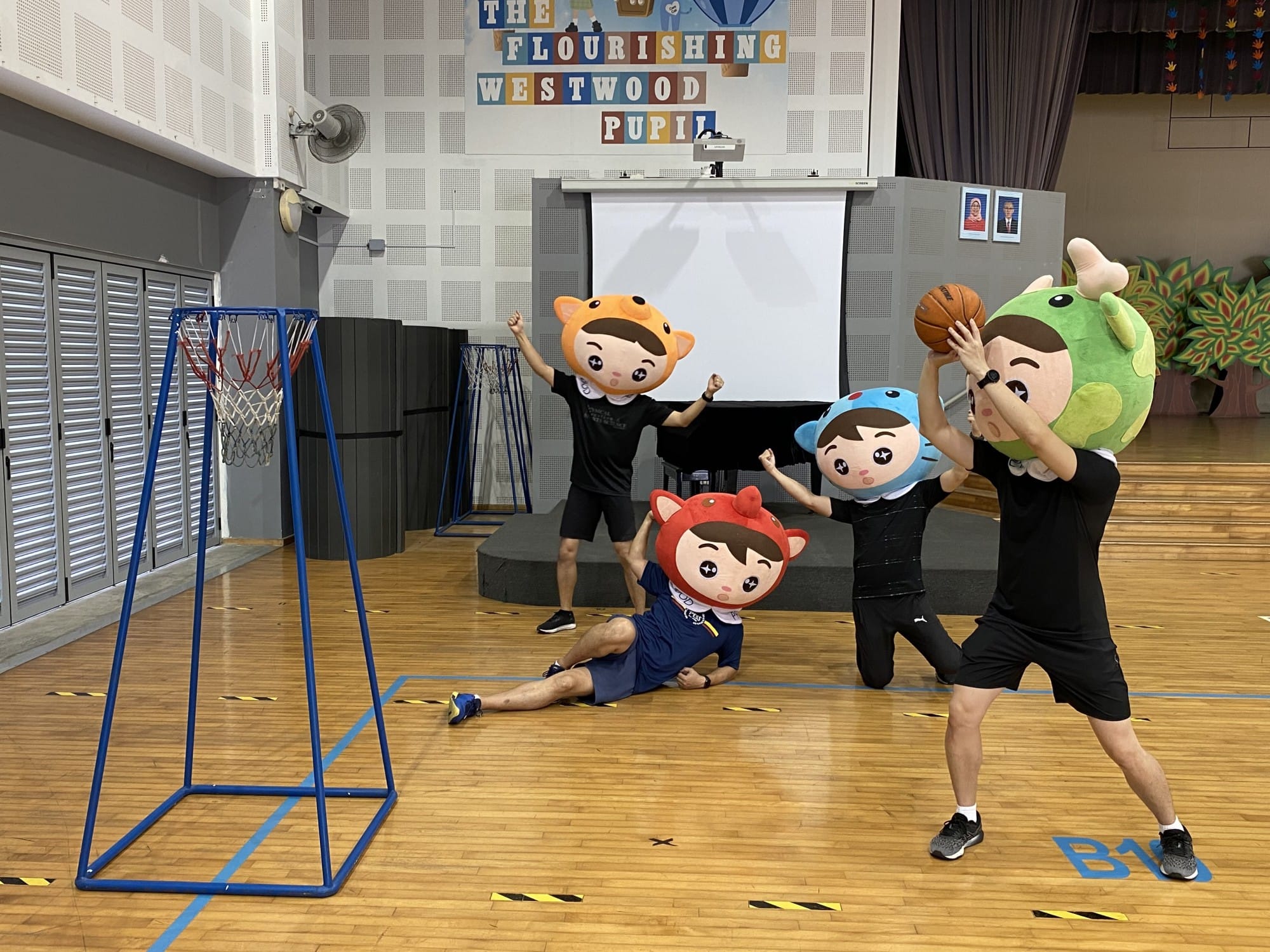 Four people wearing oversized cartoon mascot heads play basketball in a school hall with a blue hoop stand.