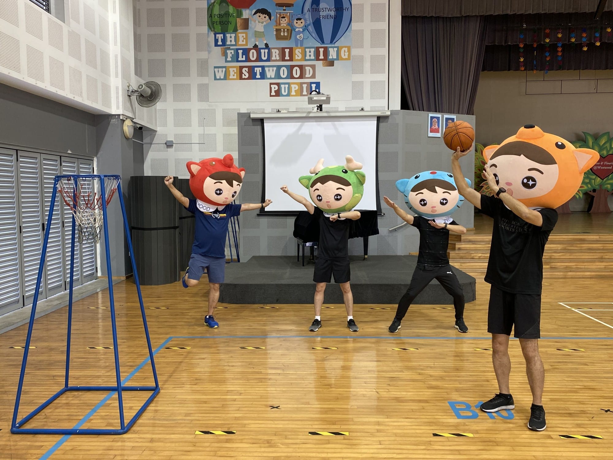 Four people wearing large cartoon mascot heads play basketball in a school hall with a Flourishing Westwood Pupil banner.
