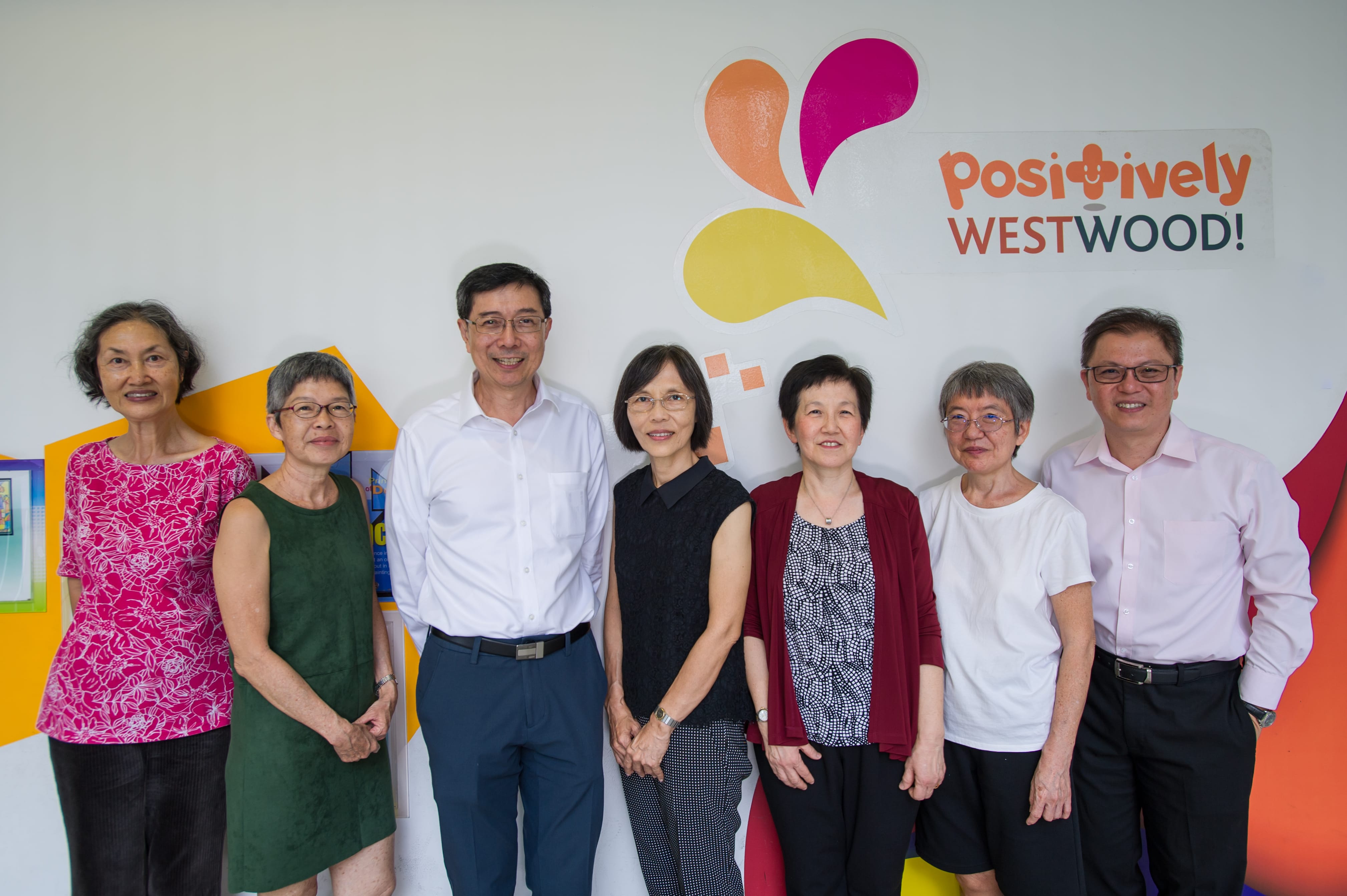 Seven adults stand smiling under a Positively Westwood sign with colorful shapes on a white wall.