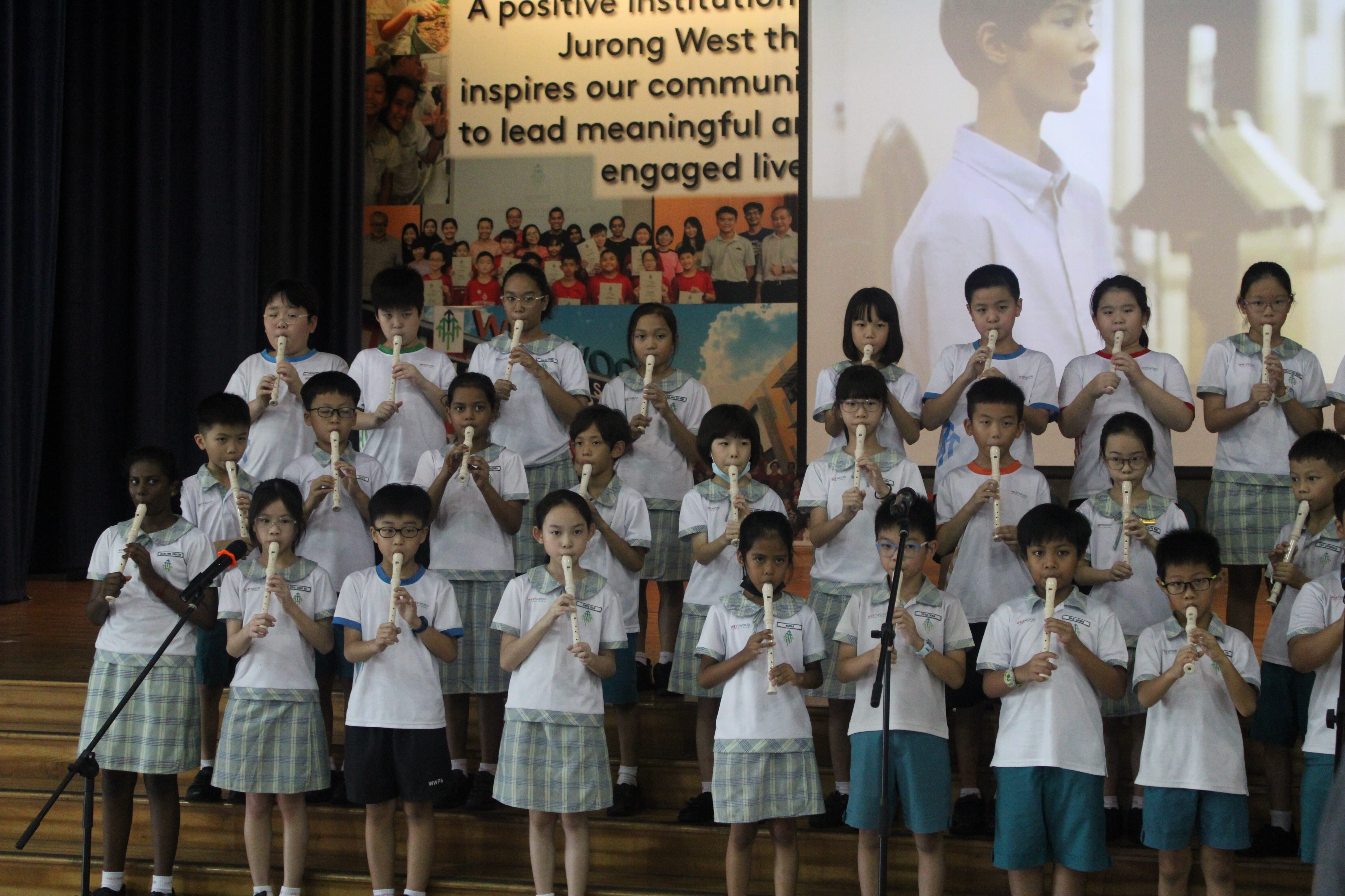 Schoolchildren in uniform stand on stage playing recorders, with microphones and a large projected singer behind.