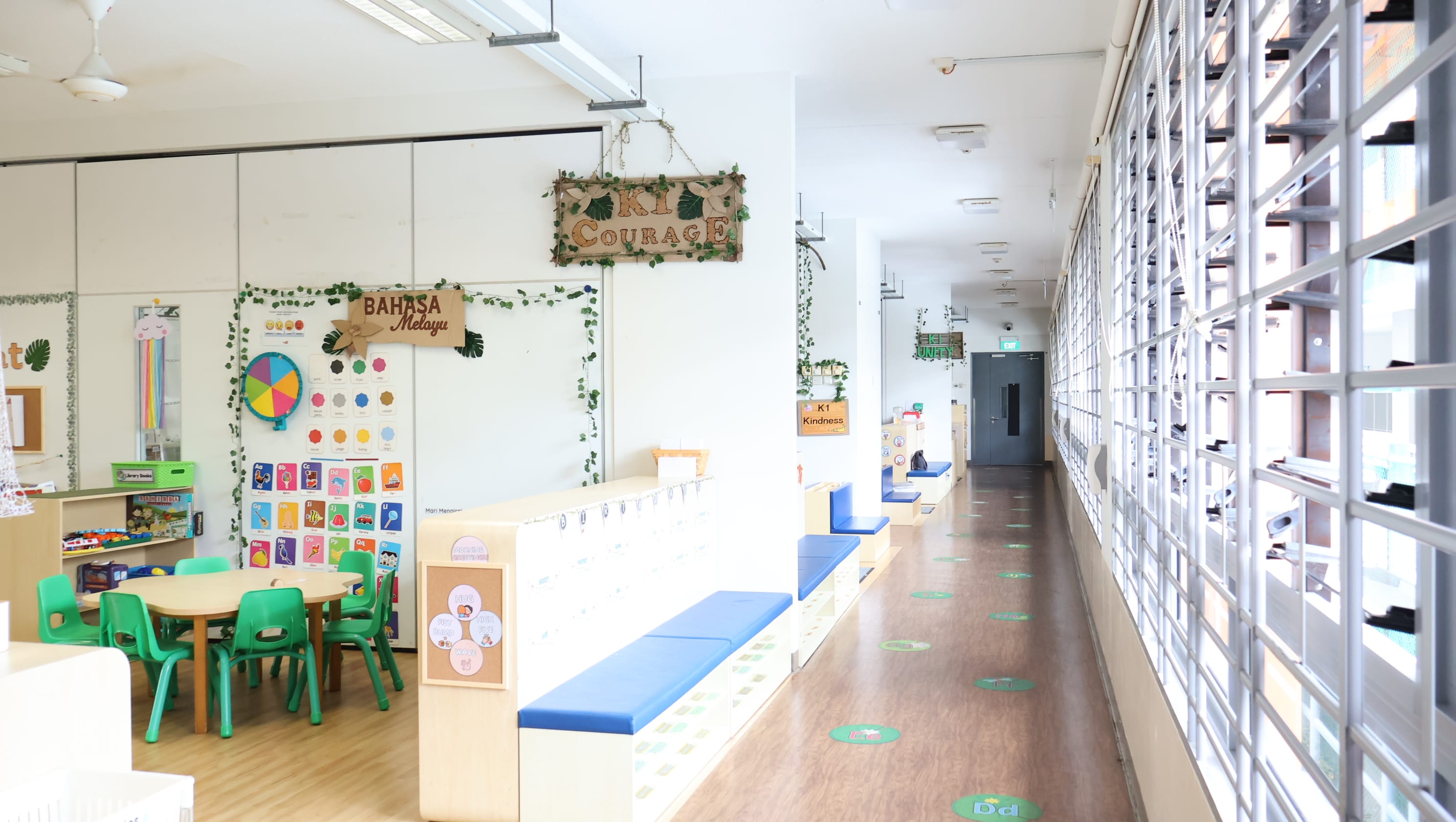 Bright kindergarten hallway with a K1 Courage sign, Bahasa Melayu board, green chairs, and blue bench seating.