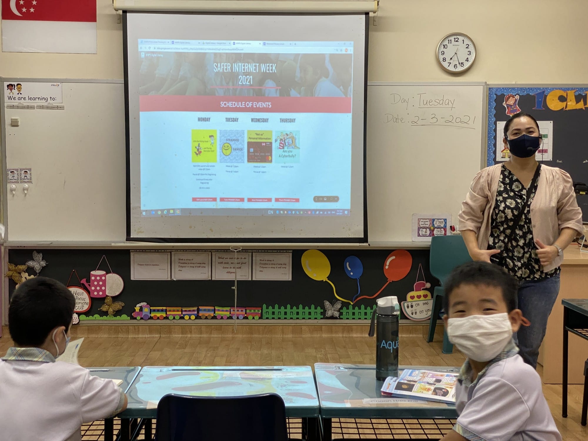 A masked teacher stands near a projector showing Safer Internet Week 2021 slides, with two masked young students seated at desks.