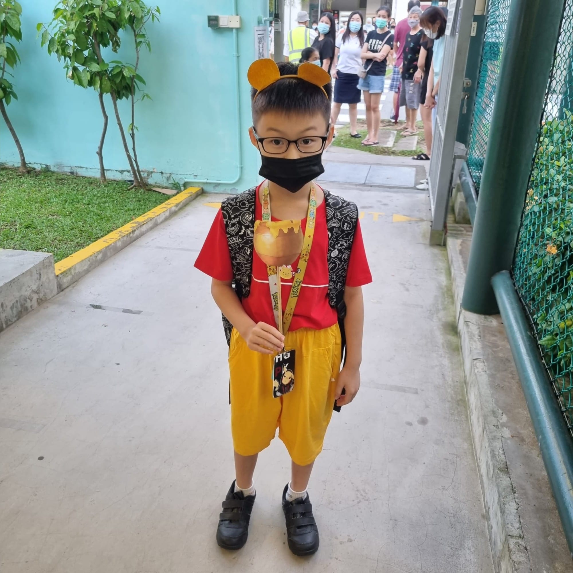 Young boy in Winnie the Pooh costume with bear ears headband, black mask, glasses, and backpack standing on a sidewalk.