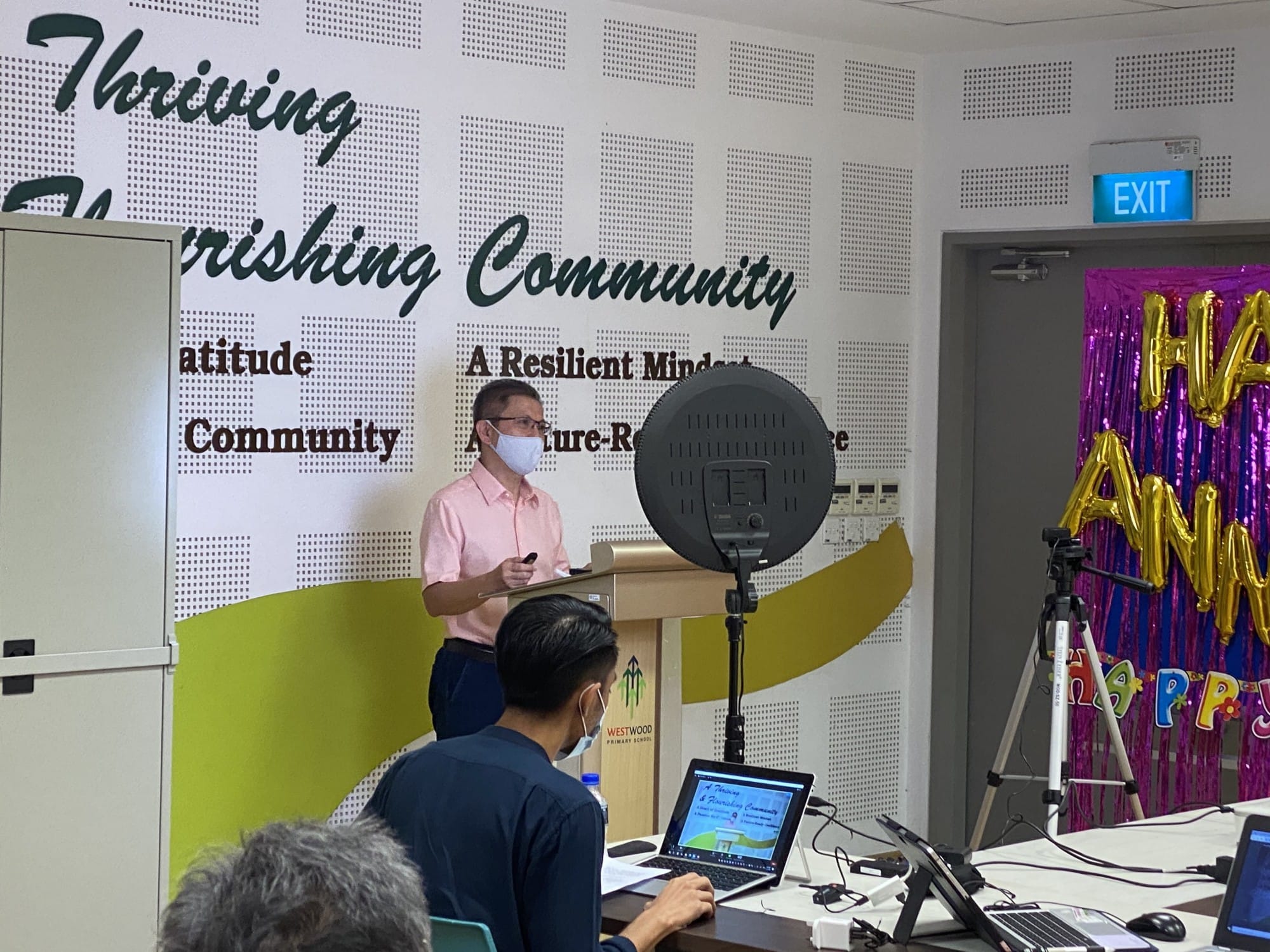 A masked man speaks at a podium before a Westwood Primary School "Thriving Community" wall mural.