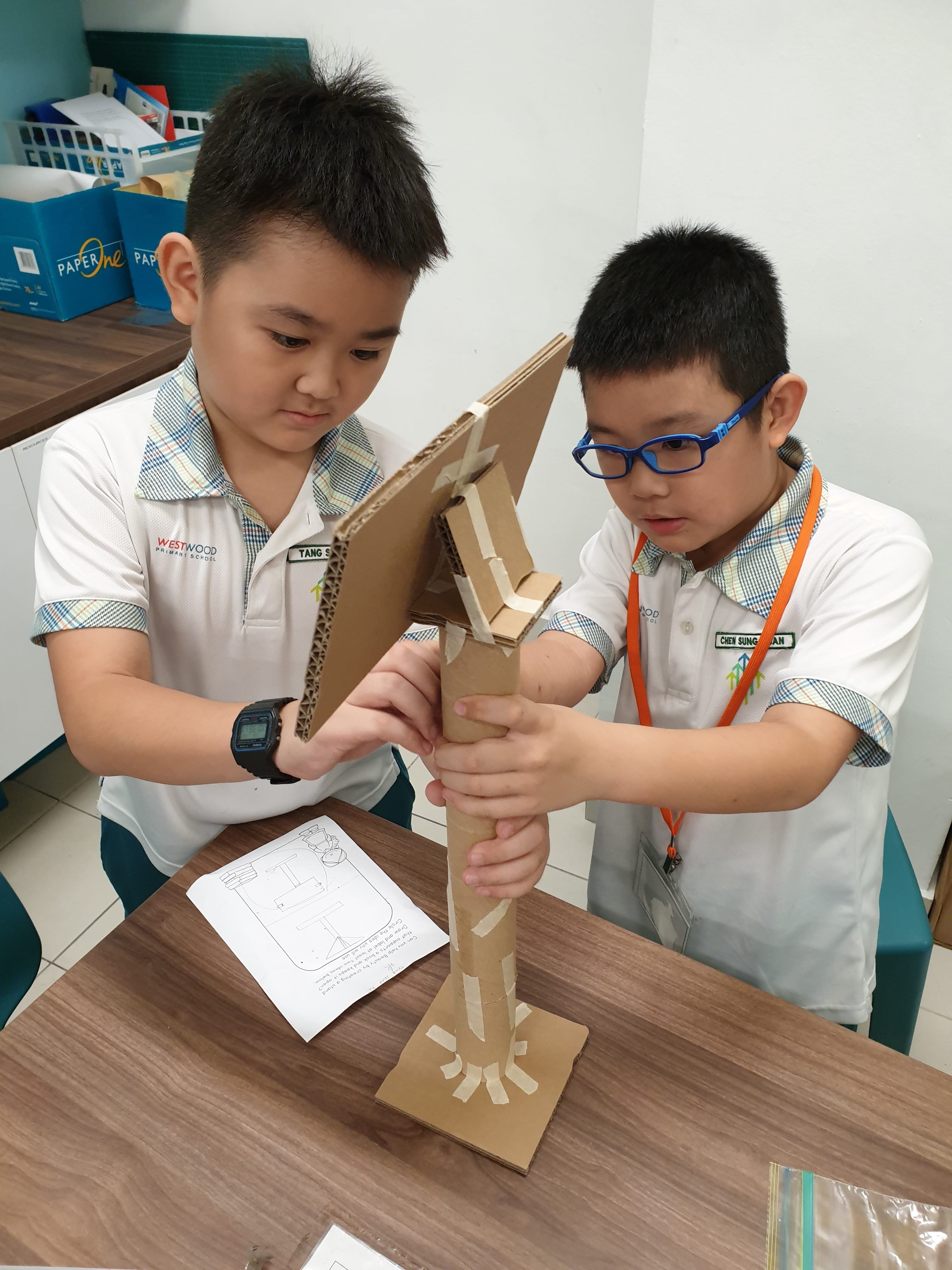Two young boys in school uniforms assembling a cardboard structure on a desk in a classroom.