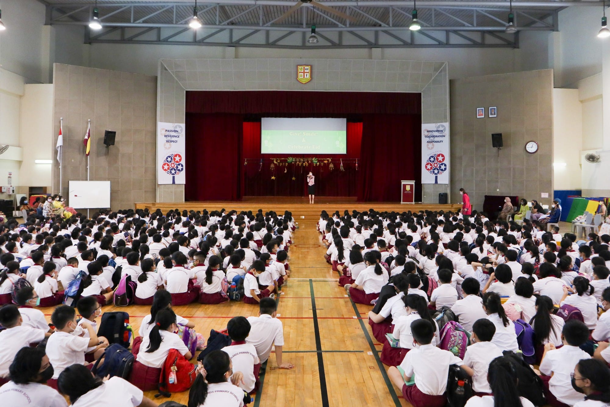 Hundreds of uniformed primary school students sit on a gymnasium floor facing a speaker on a stage with a projected screen.