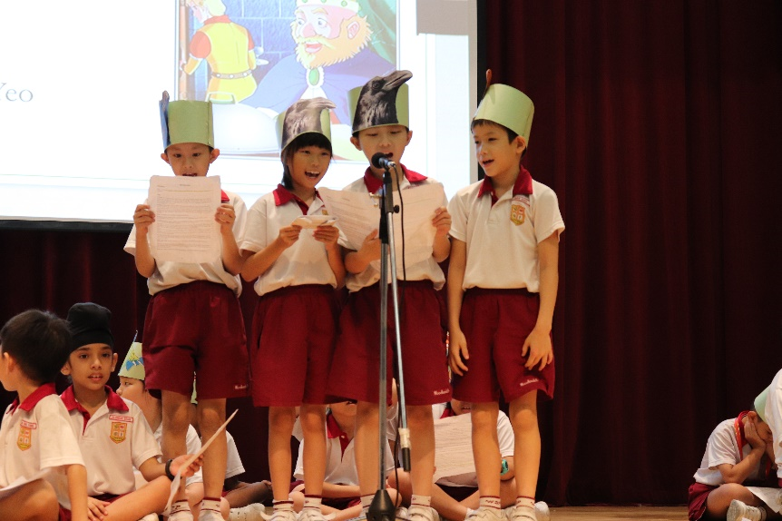 Four young students in school uniforms and paper hats perform at a microphone on stage, reading from papers.