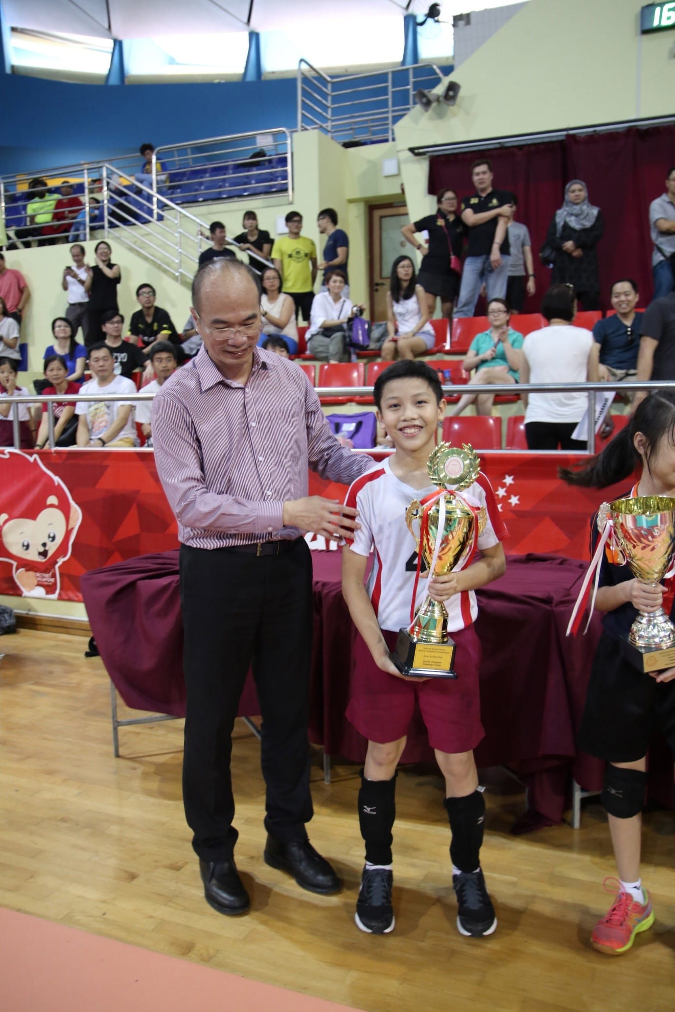 An adult man places his hand on a smiling boy holding a gold trophy at an indoor sports award ceremony.