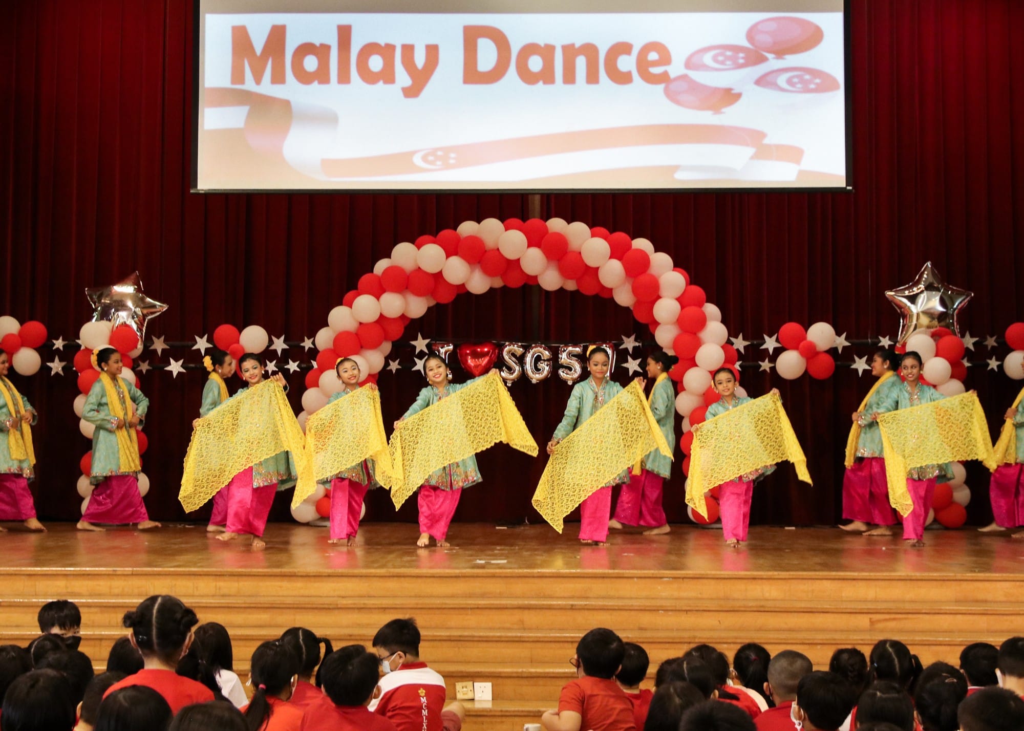 Girls in teal and pink costumes perform Malay Dance on stage holding yellow lace scarves under a red balloon arch.