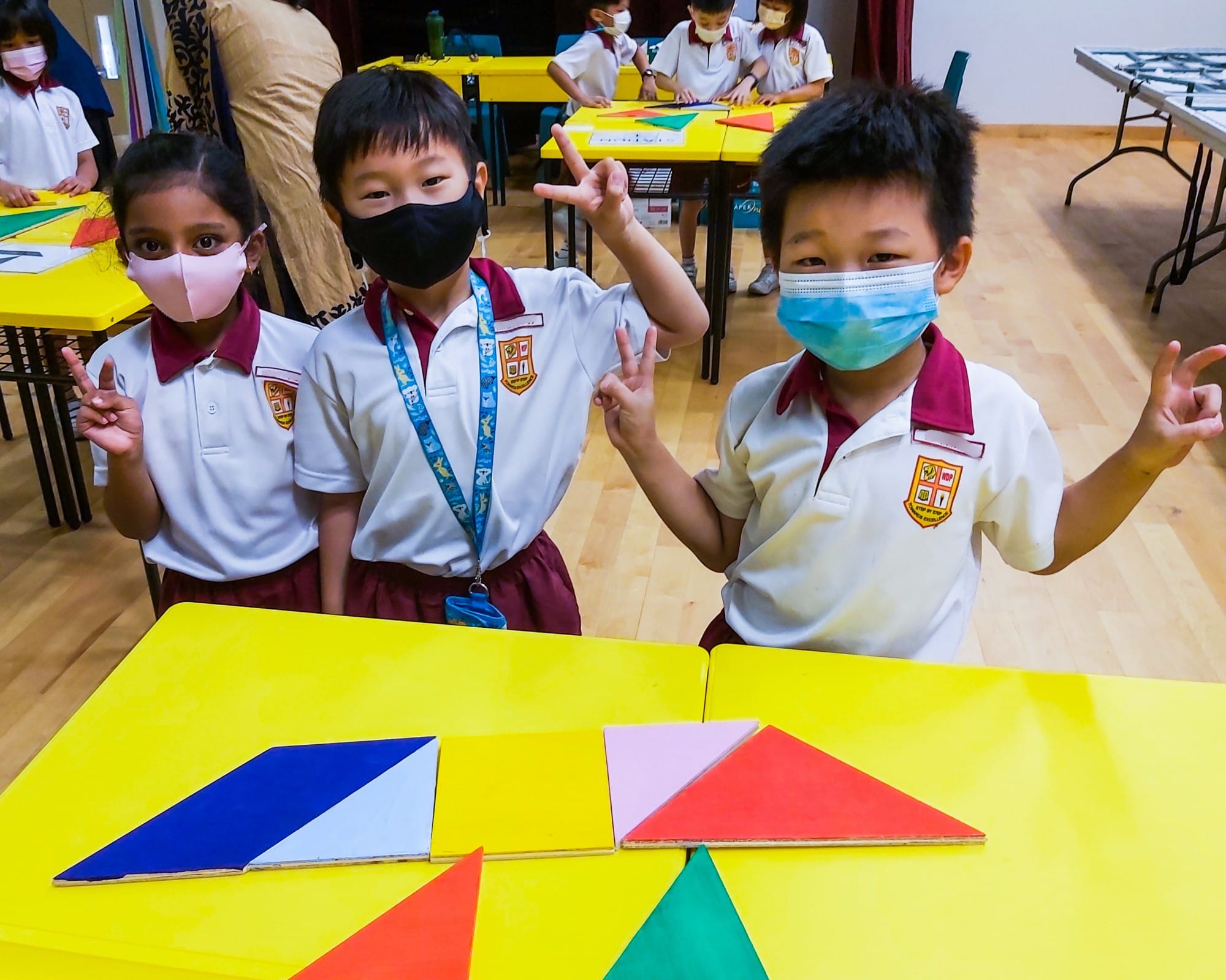 Three masked young students in school uniforms pose at a yellow table with colorful geometric shape pieces.