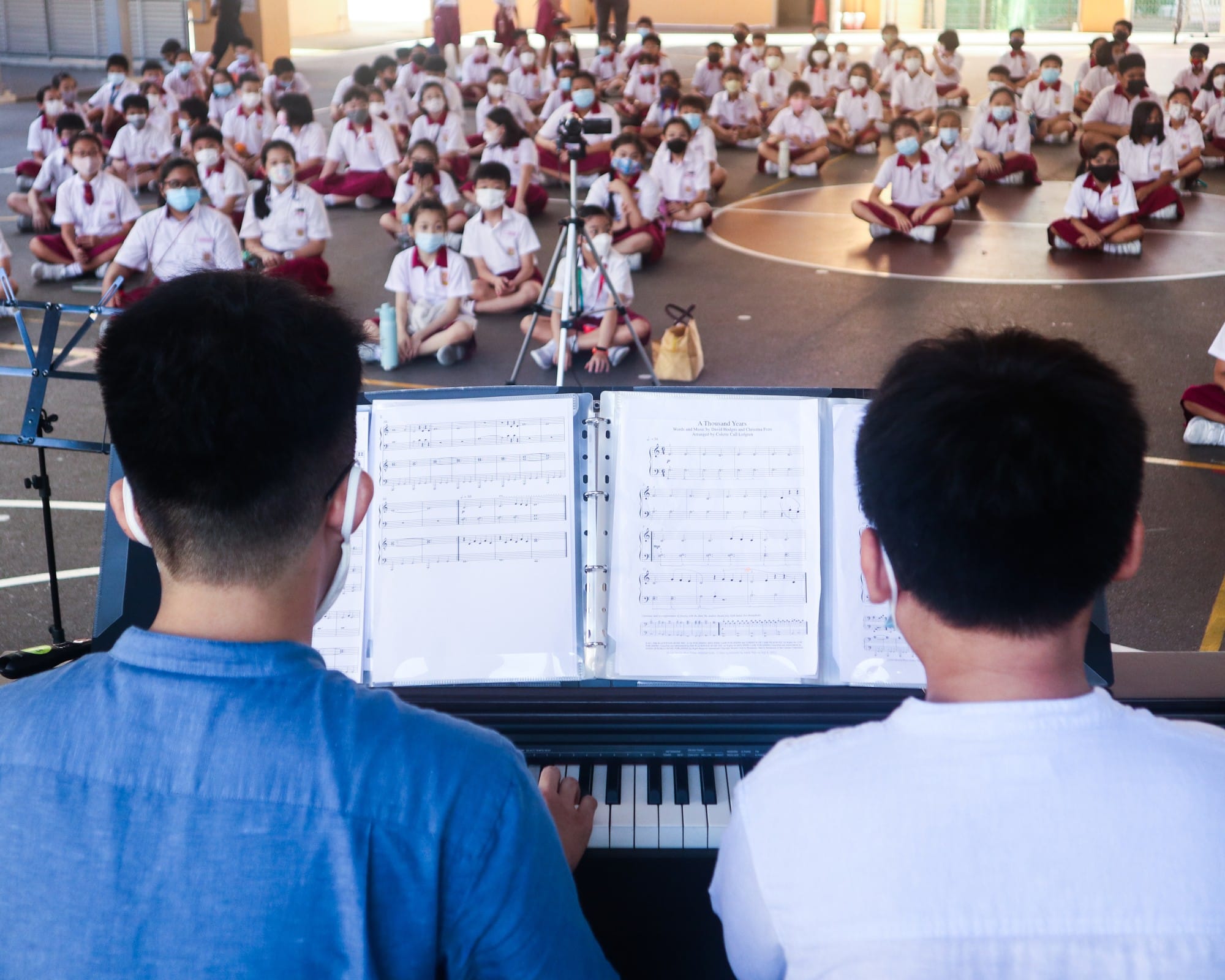 Two people at a piano with sheet music perform for a large group of masked school children sitting on a courtyard floor.