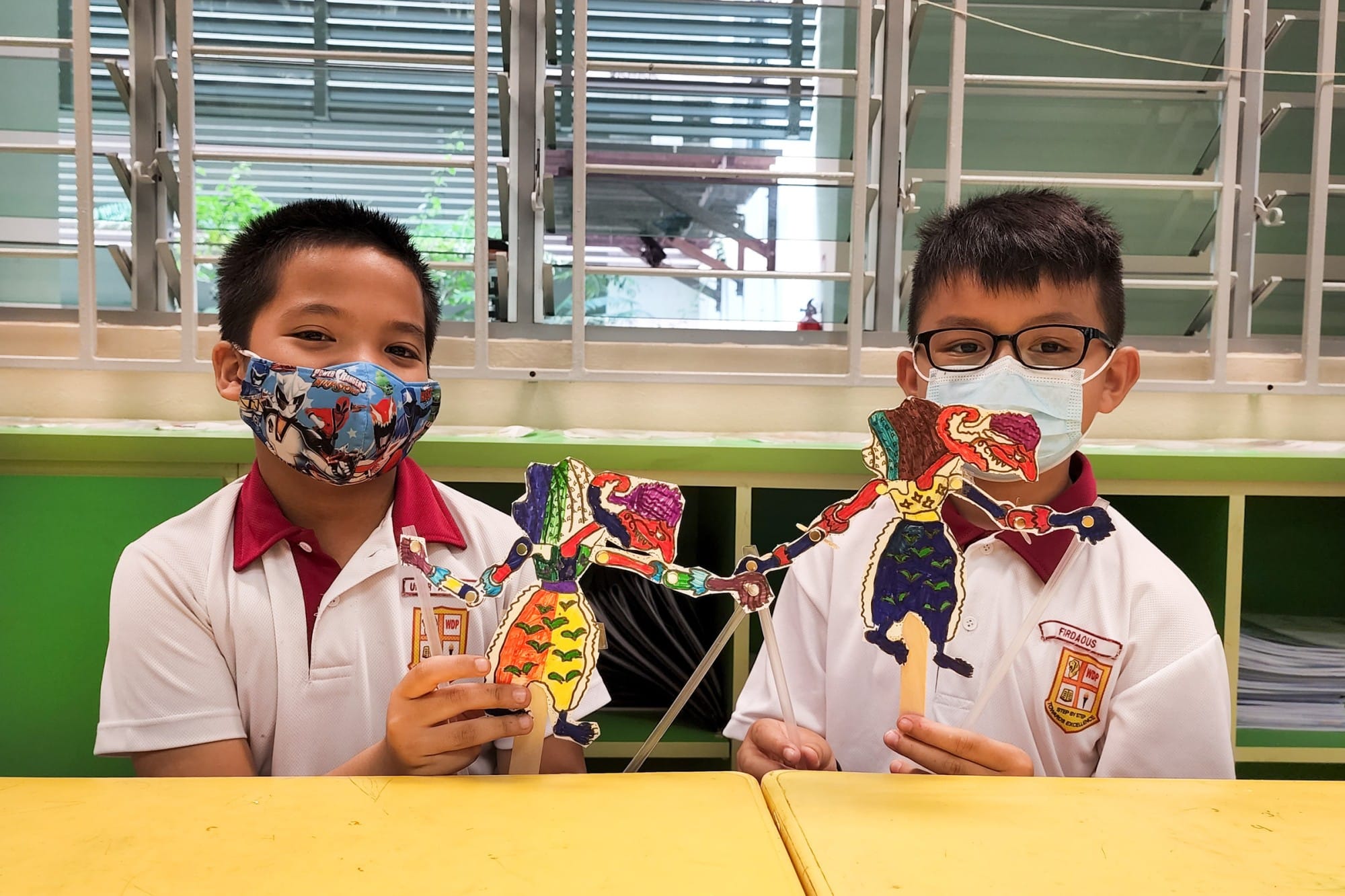 Two masked school boys hold colorful handmade puppet figures on sticks at a yellow table in a classroom.