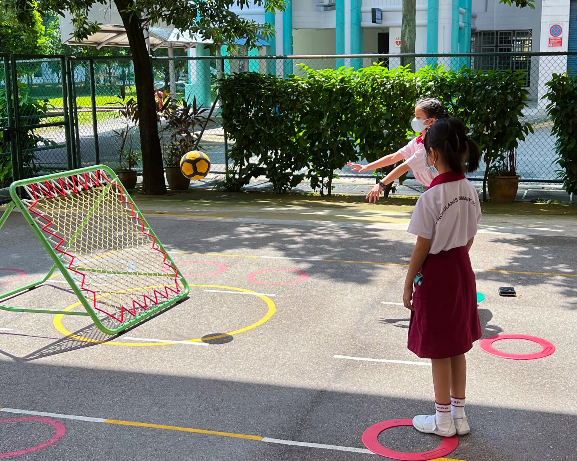 Two school girls in uniforms and masks play with a yellow ball and a green rebound net on an outdoor court.