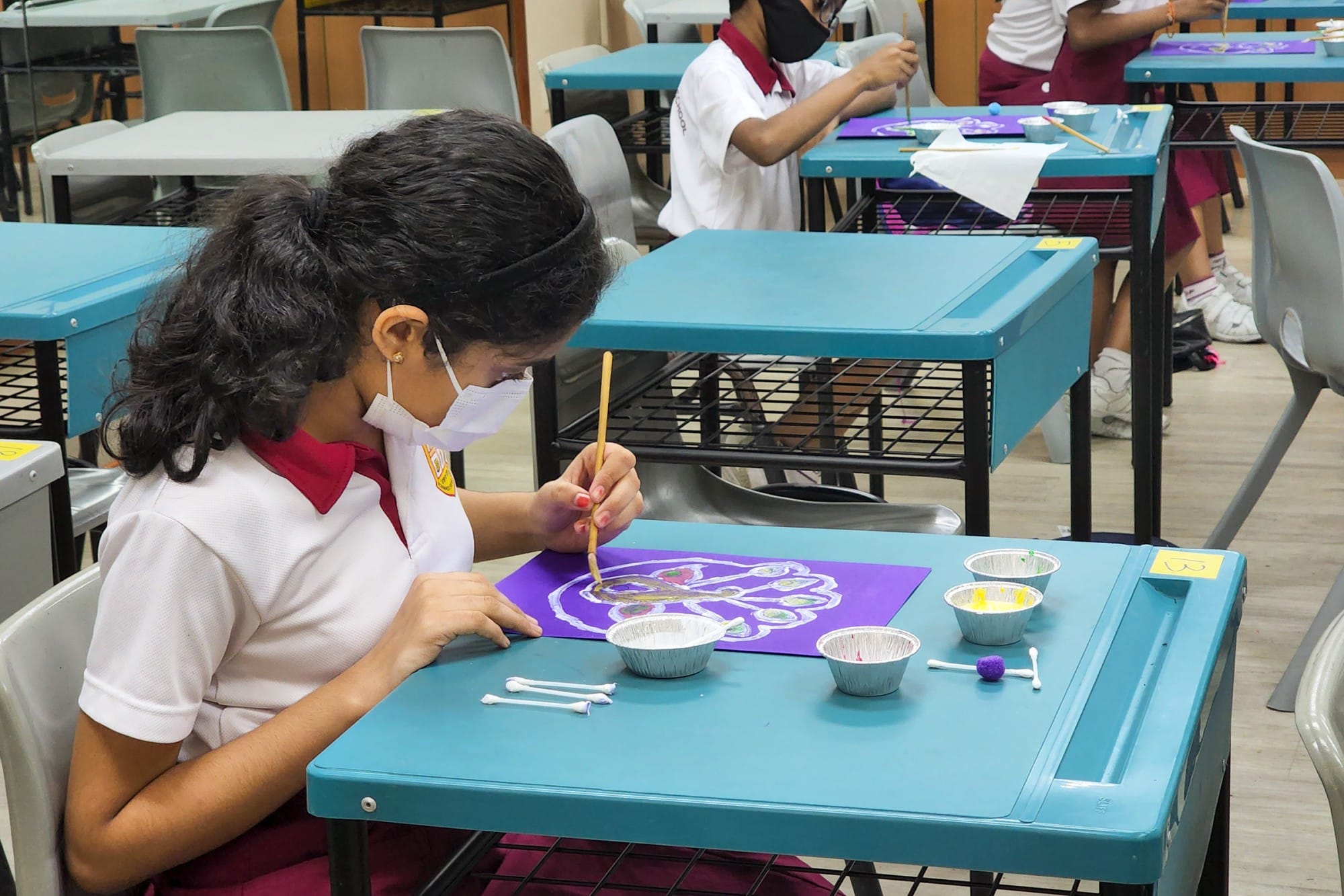 A masked schoolgirl in uniform paints on purple paper at a teal desk with small paint cups nearby.