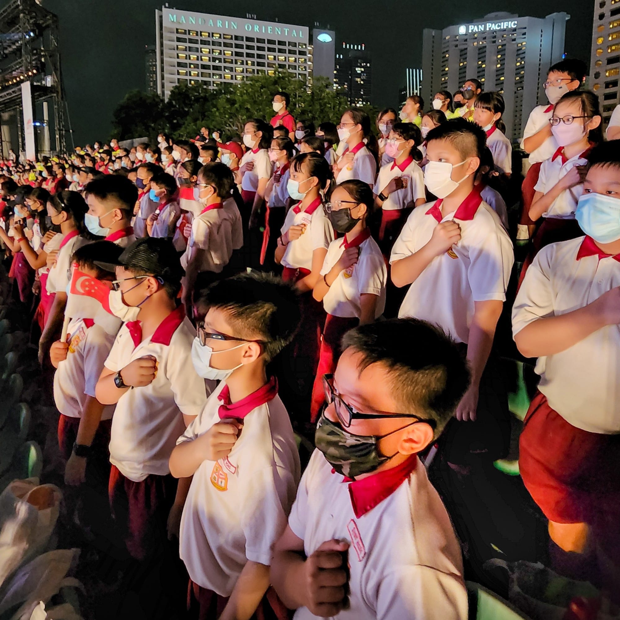 Large crowd of masked schoolchildren in white and red uniforms standing at attention outdoors at night near city buildings.