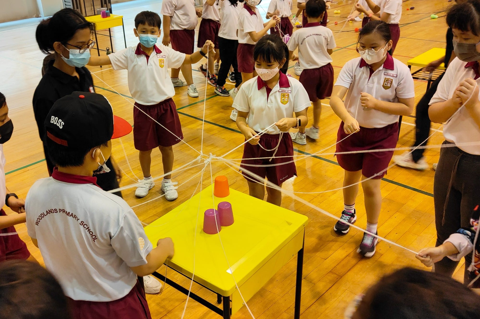 Children in school uniforms and masks use strings attached to a rubber band to stack cups on a yellow table in a gym.