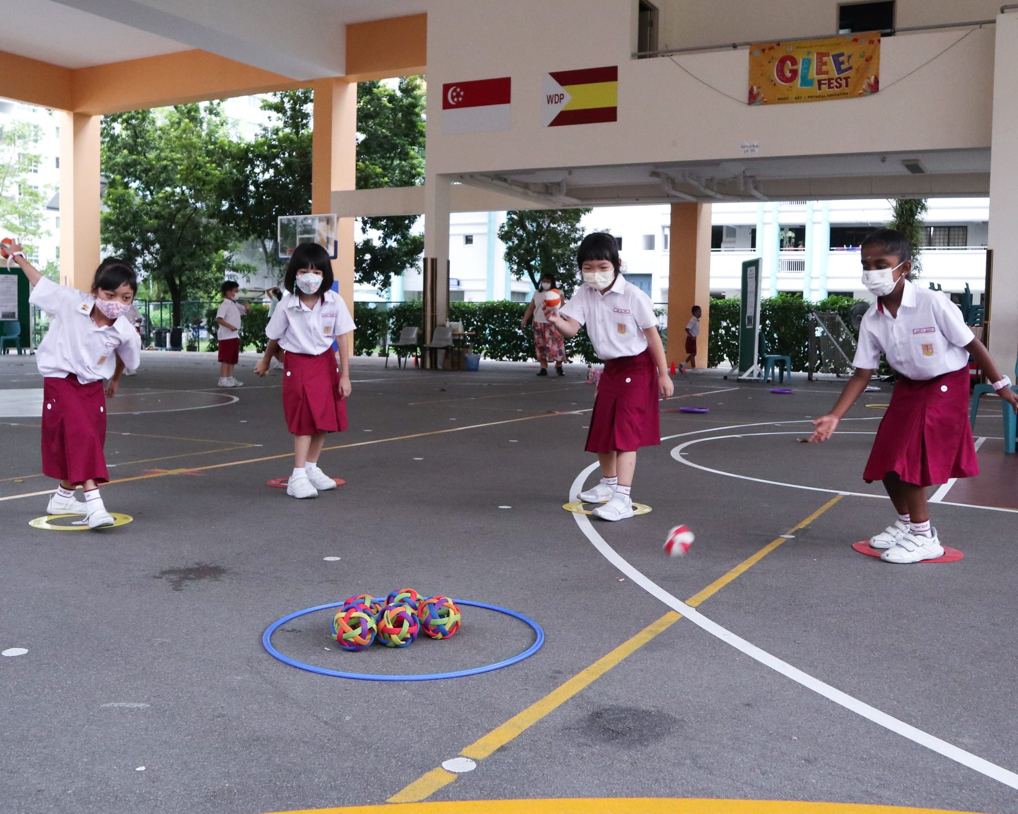 Young school girls in uniforms and masks play a ball activity on a court with colorful balls in a hoop.