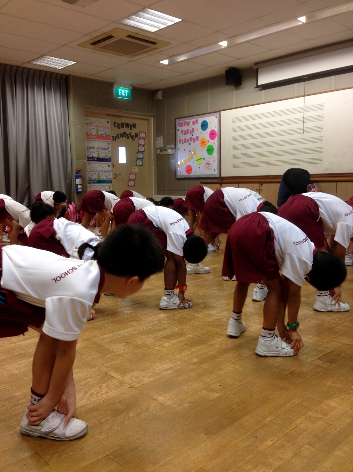 Children in white and maroon school uniforms bend forward doing stretches on a wooden floor in a school hall.