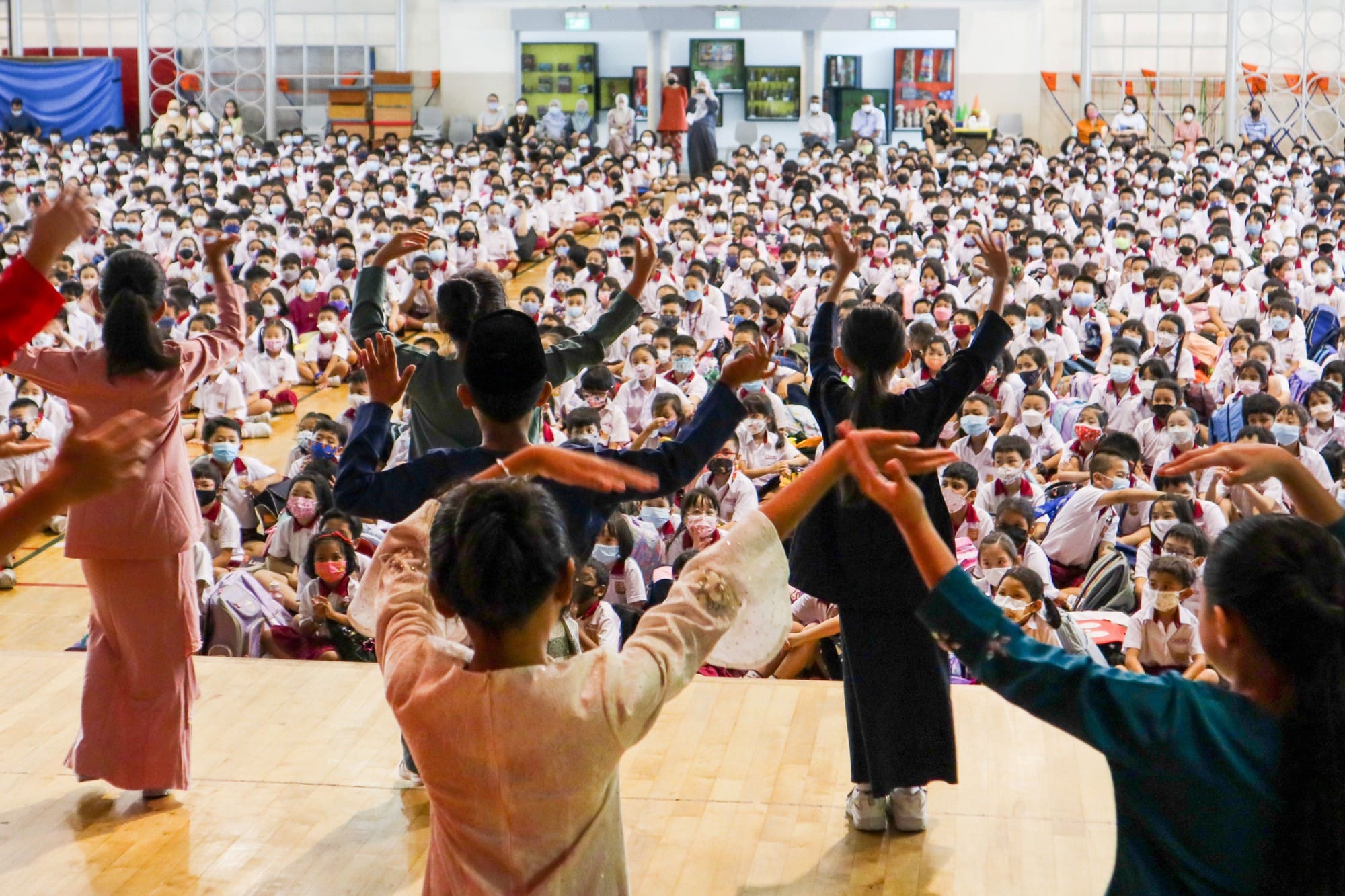 Dancers perform on a gym floor before a large crowd of masked schoolchildren seated on the ground.