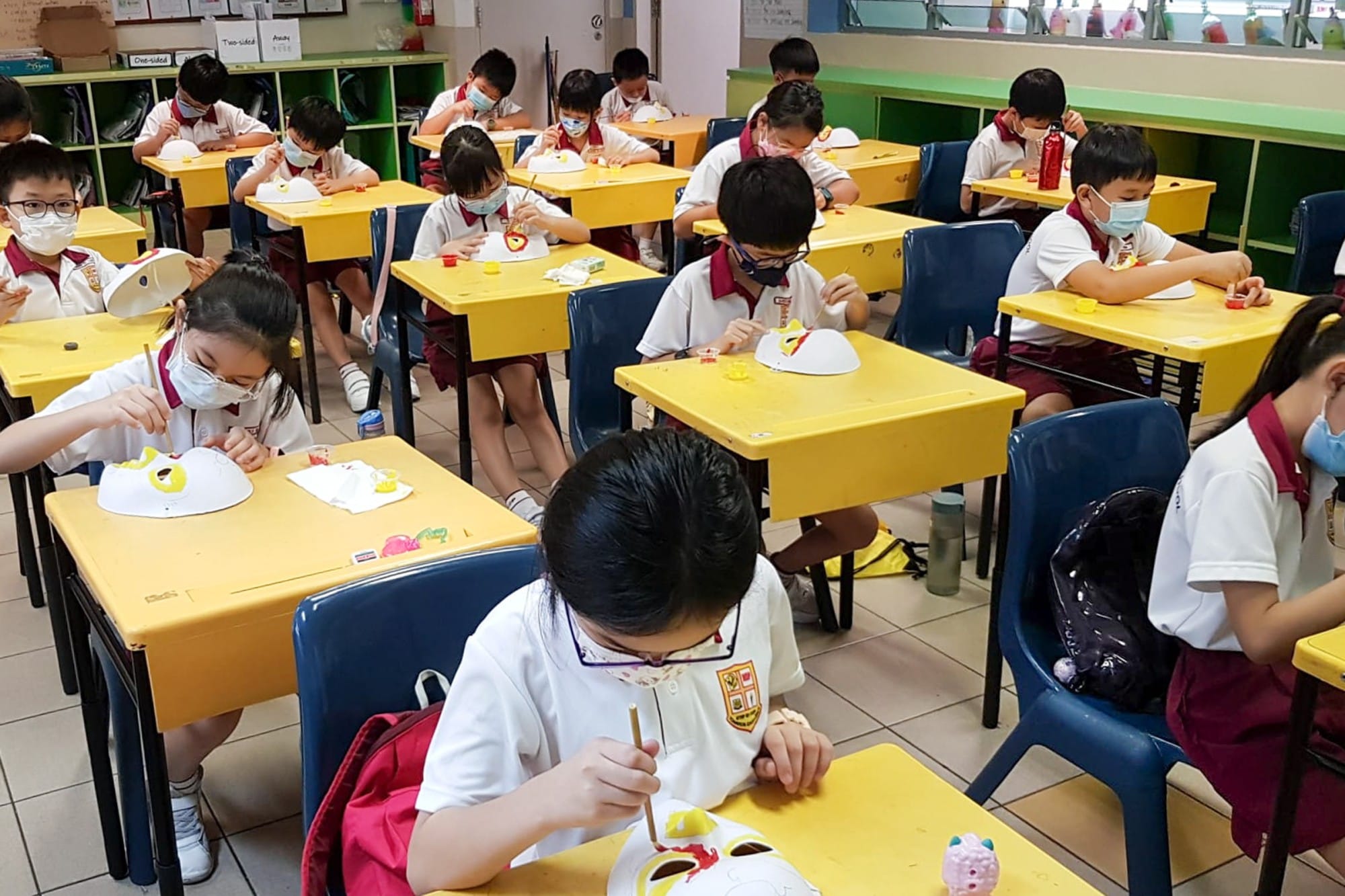 Young students wearing face masks sit at yellow desks painting white masks during an art class.