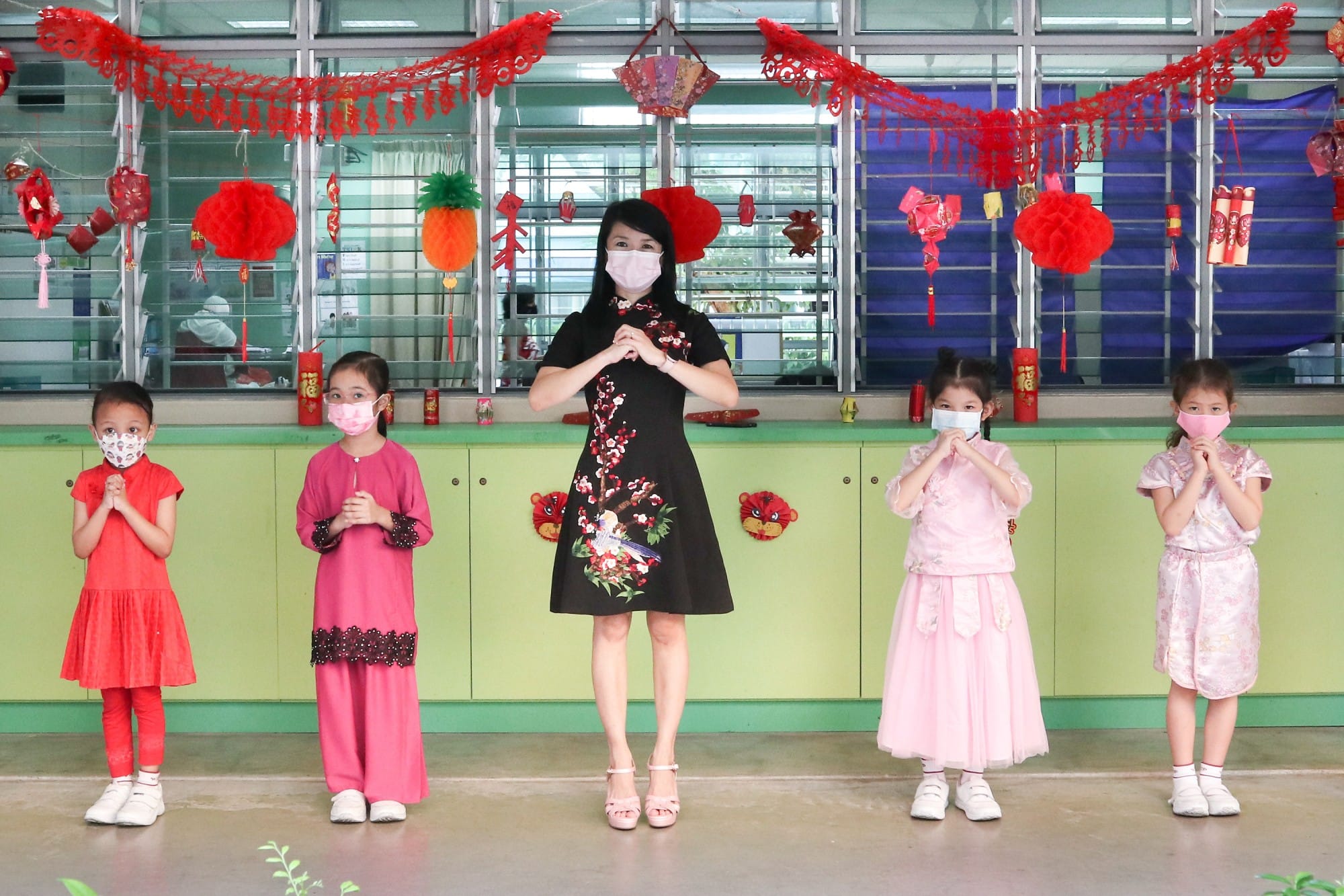 A woman and four young girls in festive attire and masks clasp hands in greeting amid red Chinese New Year decorations.