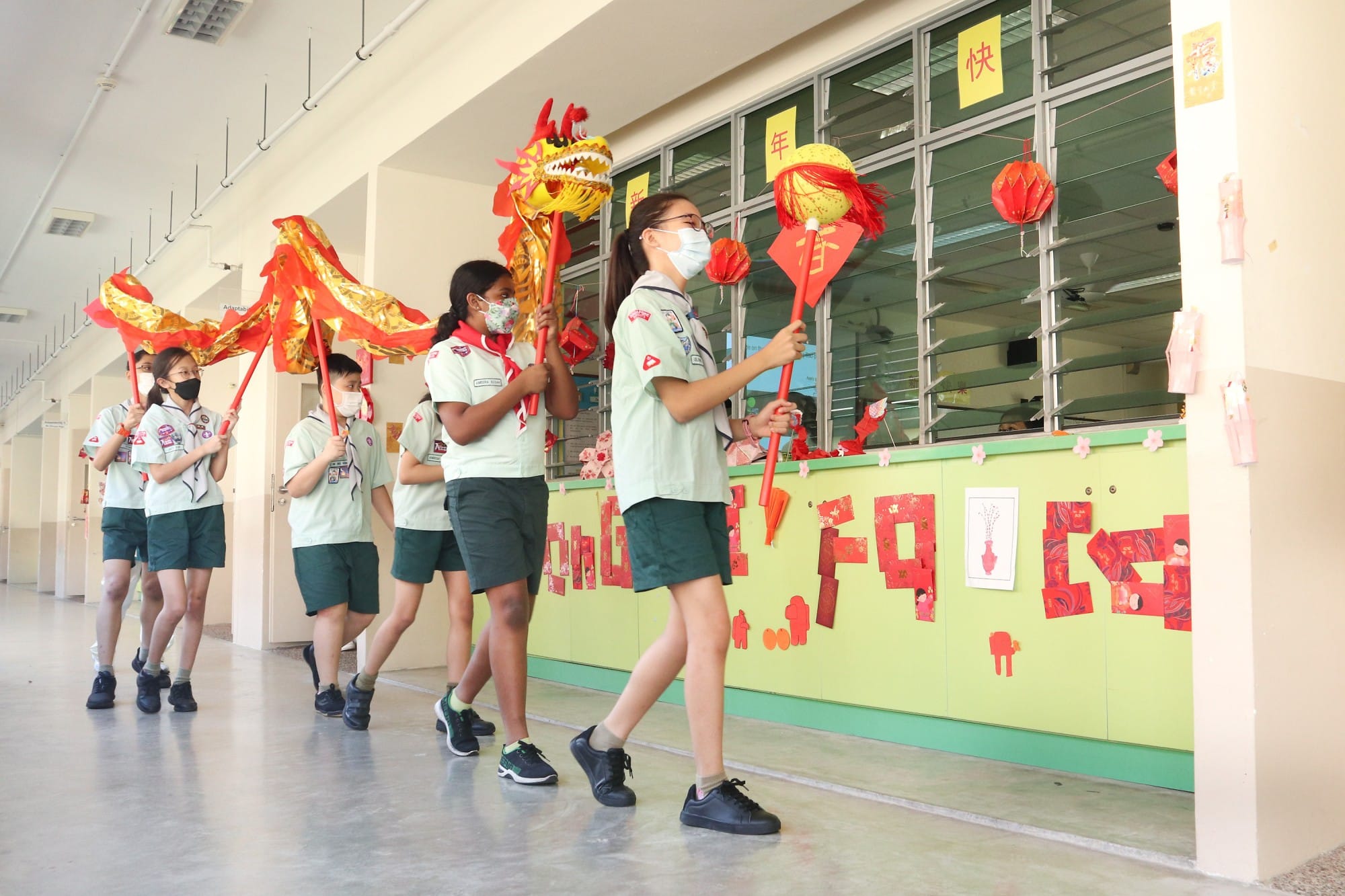 Students in scout uniforms and masks parade a red and gold dragon through a school corridor decorated for Chinese New Year.