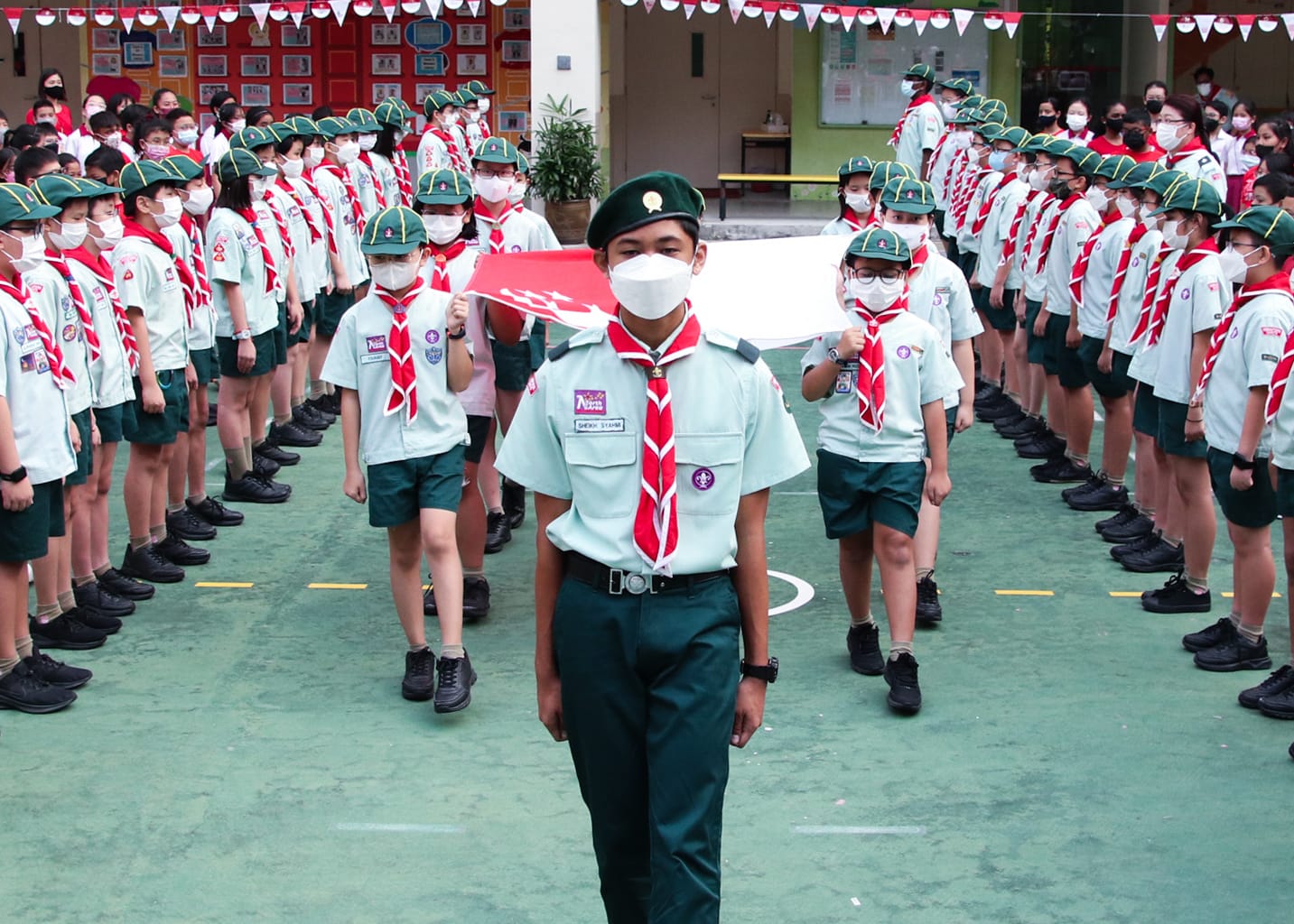 A large group of masked youth scouts in green uniforms and red neckerchiefs stand in formation outdoors.