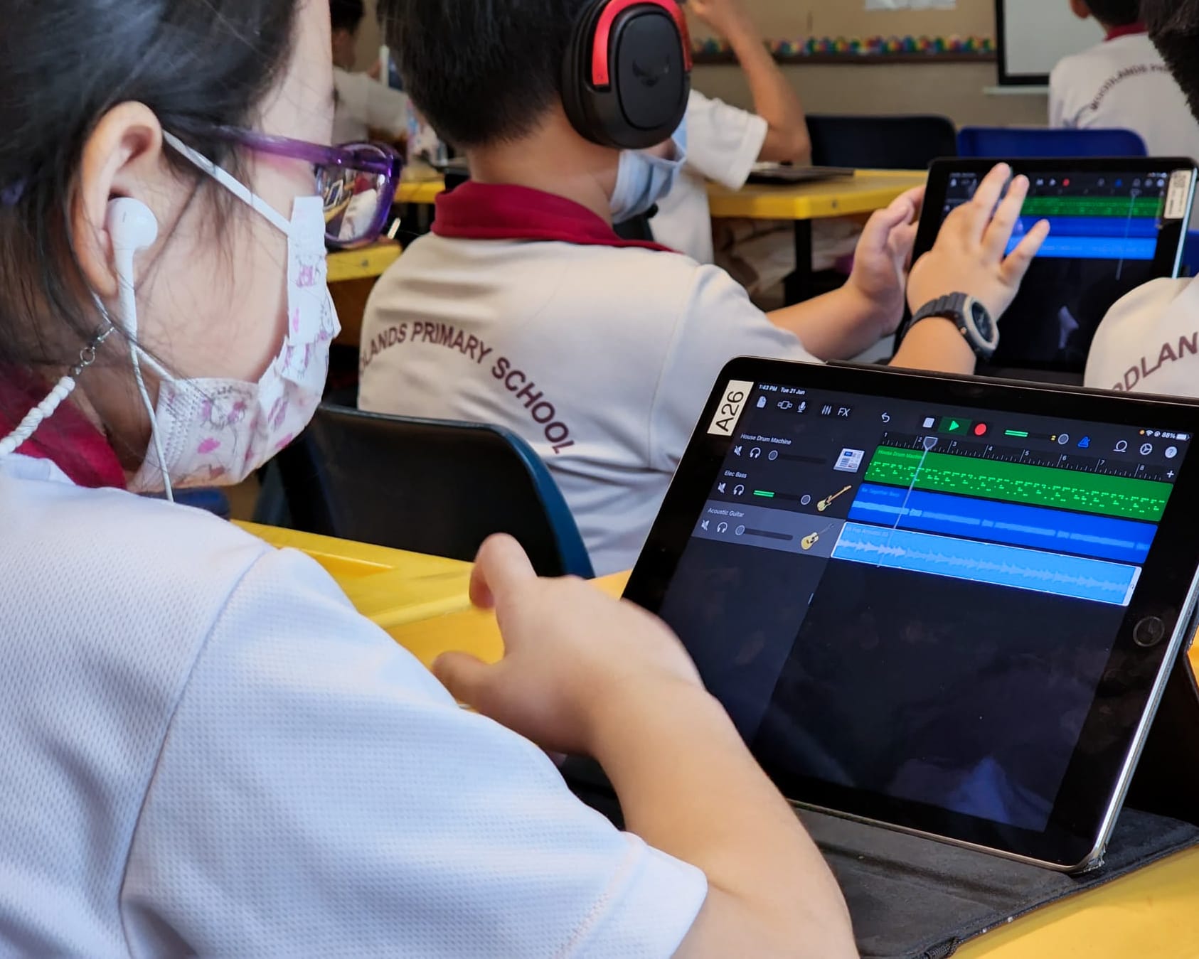 Students in school uniforms and masks use tablets running a music production app in a classroom.