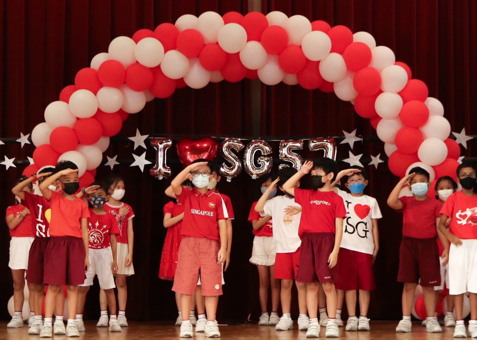 Children in red and white outfits salute on stage under a red-white balloon arch with "I Love SG57" decorations.