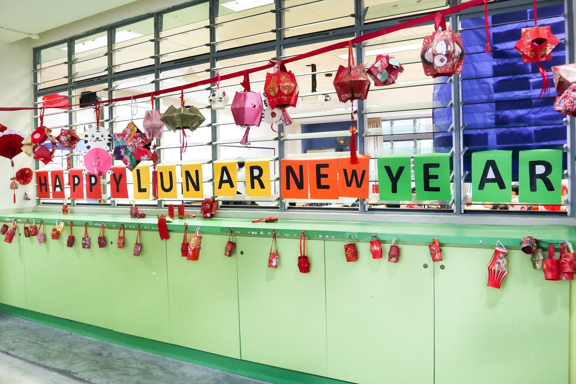 Colorful Happy Lunar New Year banner with hanging paper lanterns and red decorations in a school hallway.