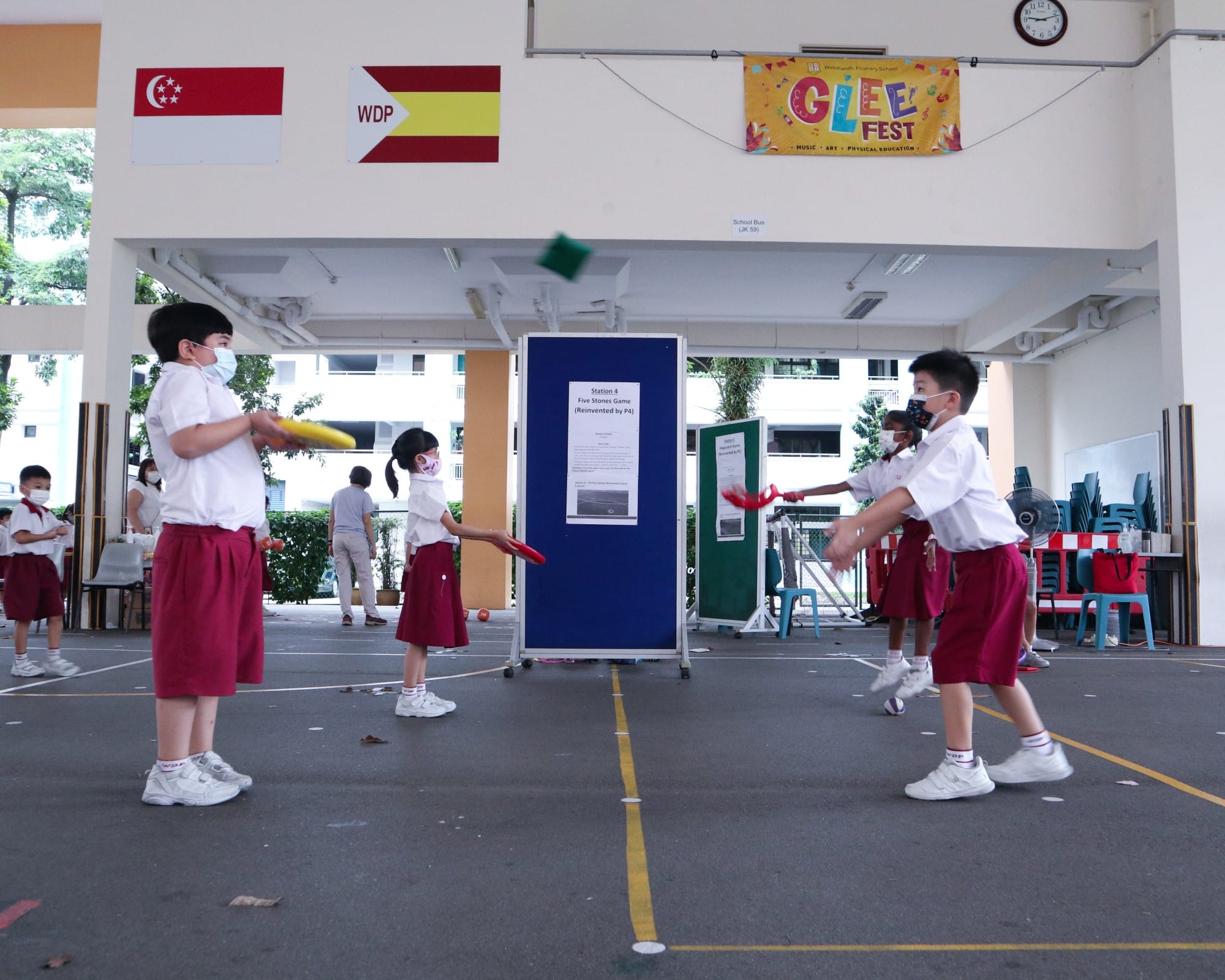 Children in school uniforms play a tossing game outdoors at a school Glee Fest event with flags on the wall.