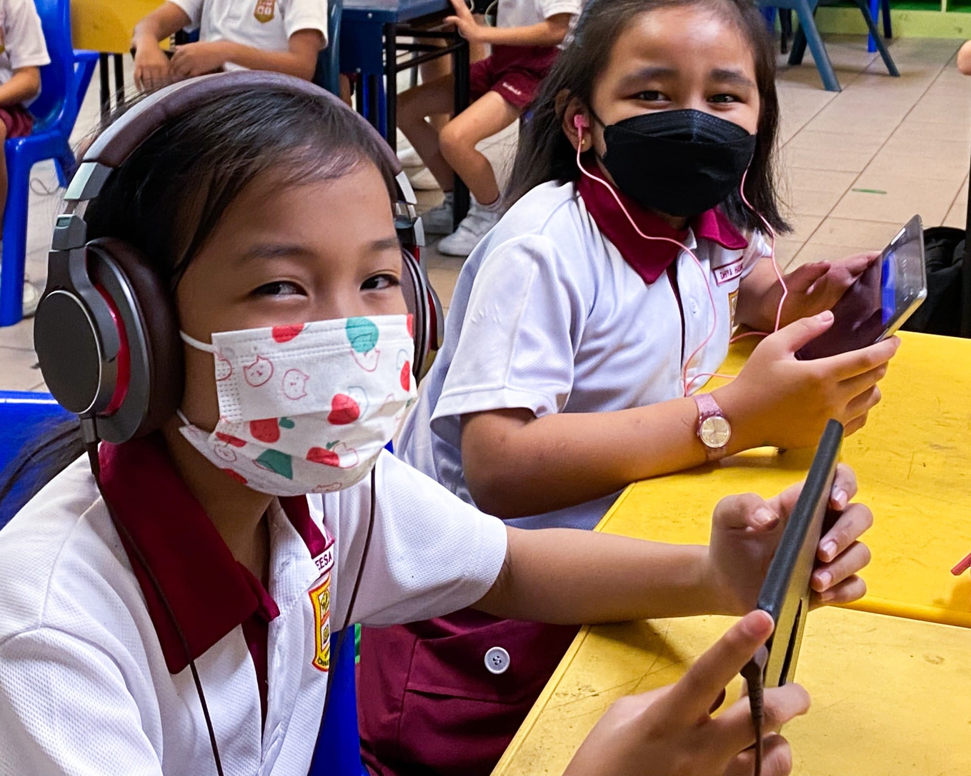 Two young girls in school uniforms and face masks use tablets, one wearing large headphones, seated at a yellow table.