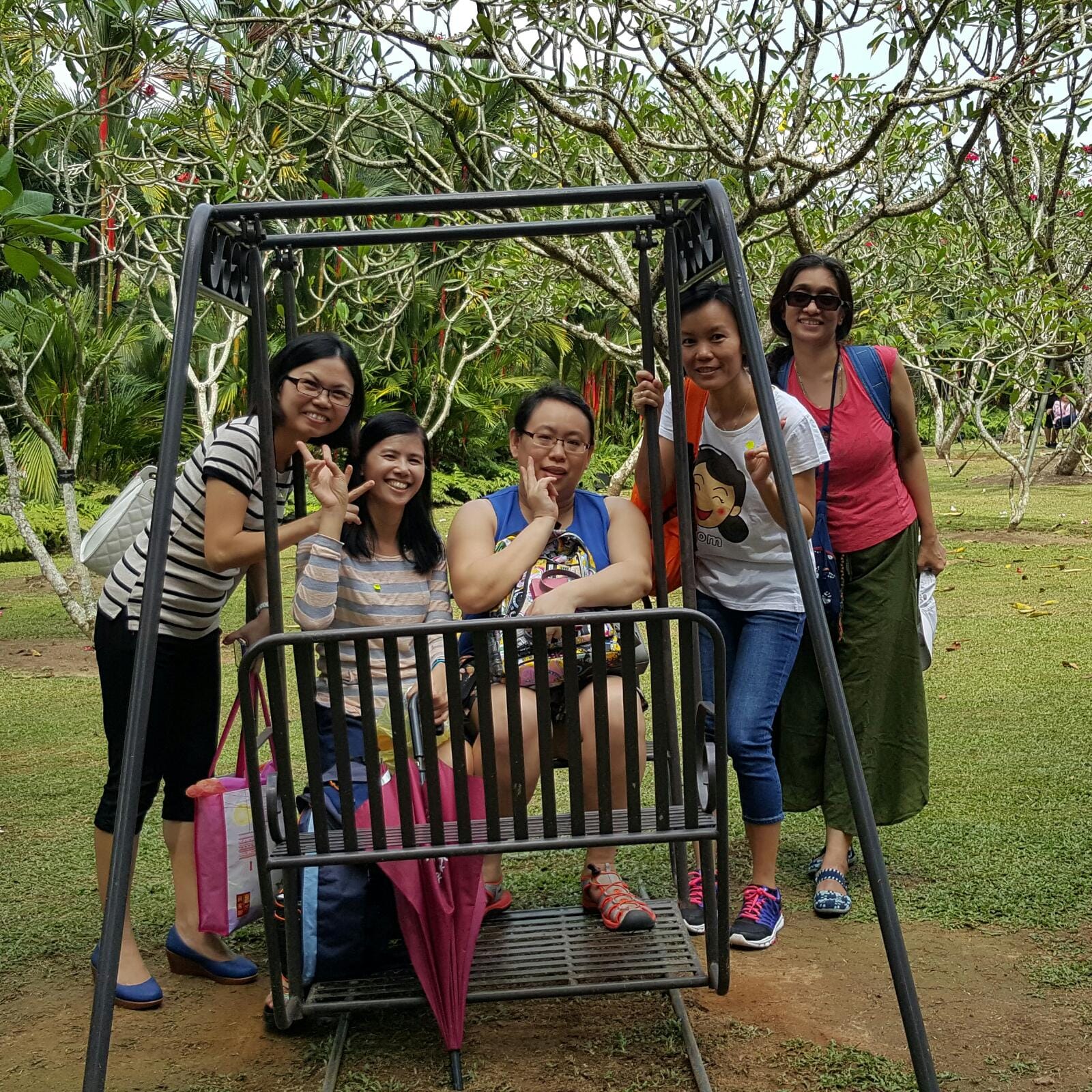 Five women posing and smiling around a metal garden swing bench surrounded by trees and green grass.