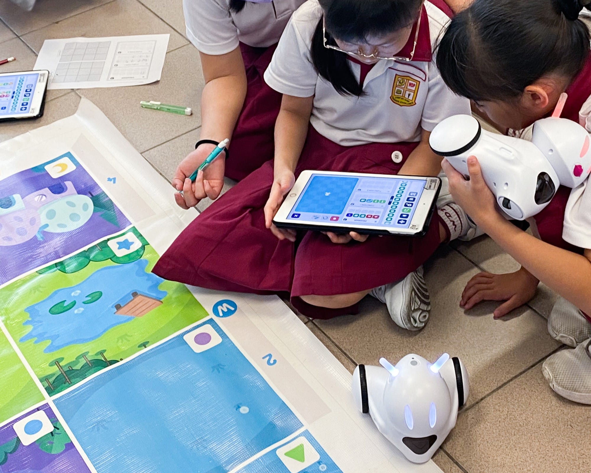 Students in school uniforms sit on a floor using a tablet to program small white robots on a colorful grid mat.
