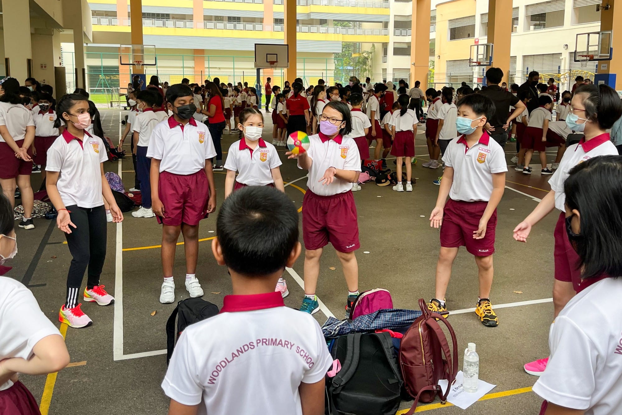 Students in Woodlands Primary School uniforms and face masks play with a colorful ball on an outdoor court.