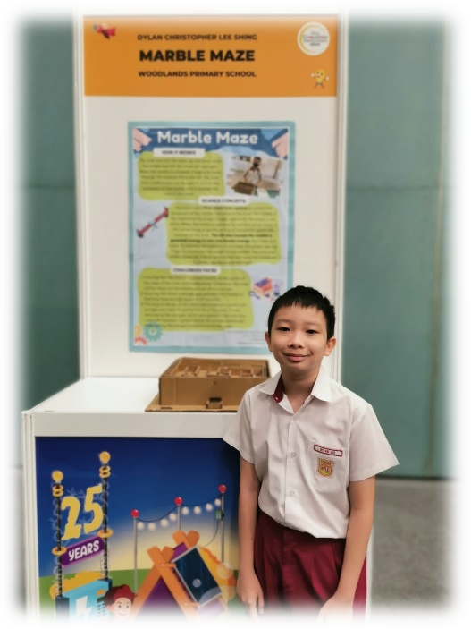 Young boy in school uniform stands beside a Marble Maze project display board from Woodlands Primary School.