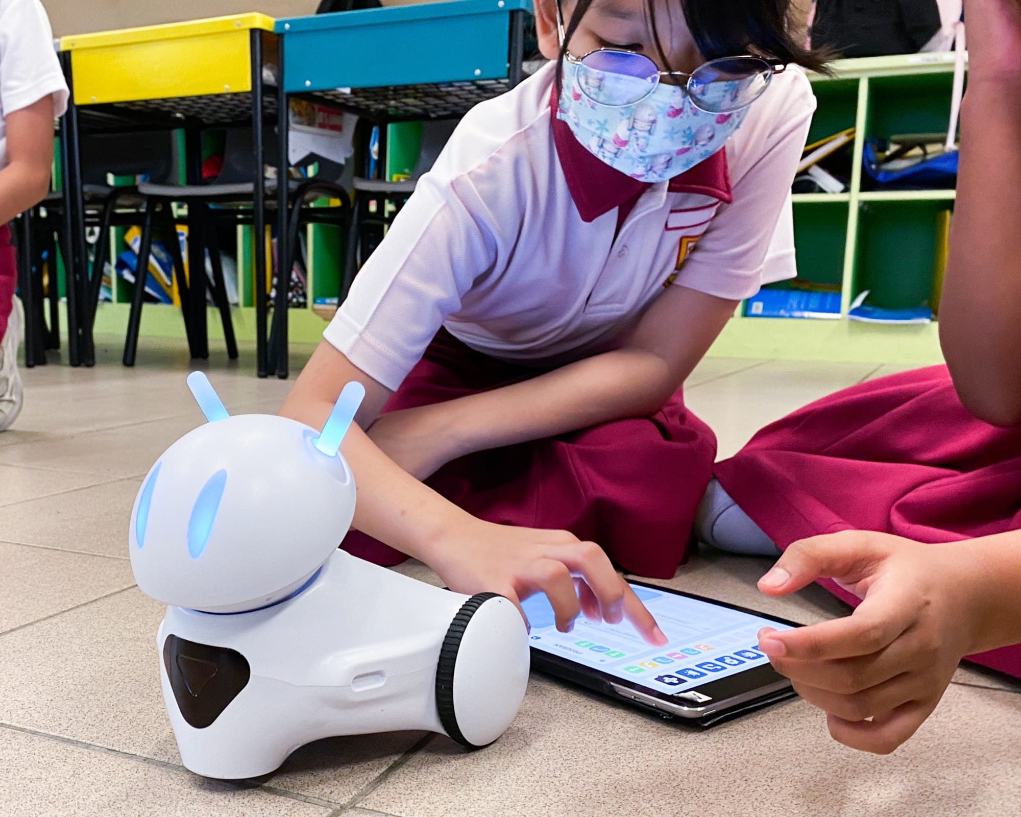 A masked schoolgirl sits on a classroom floor, tapping a tablet next to a small white robot with blue antennae.