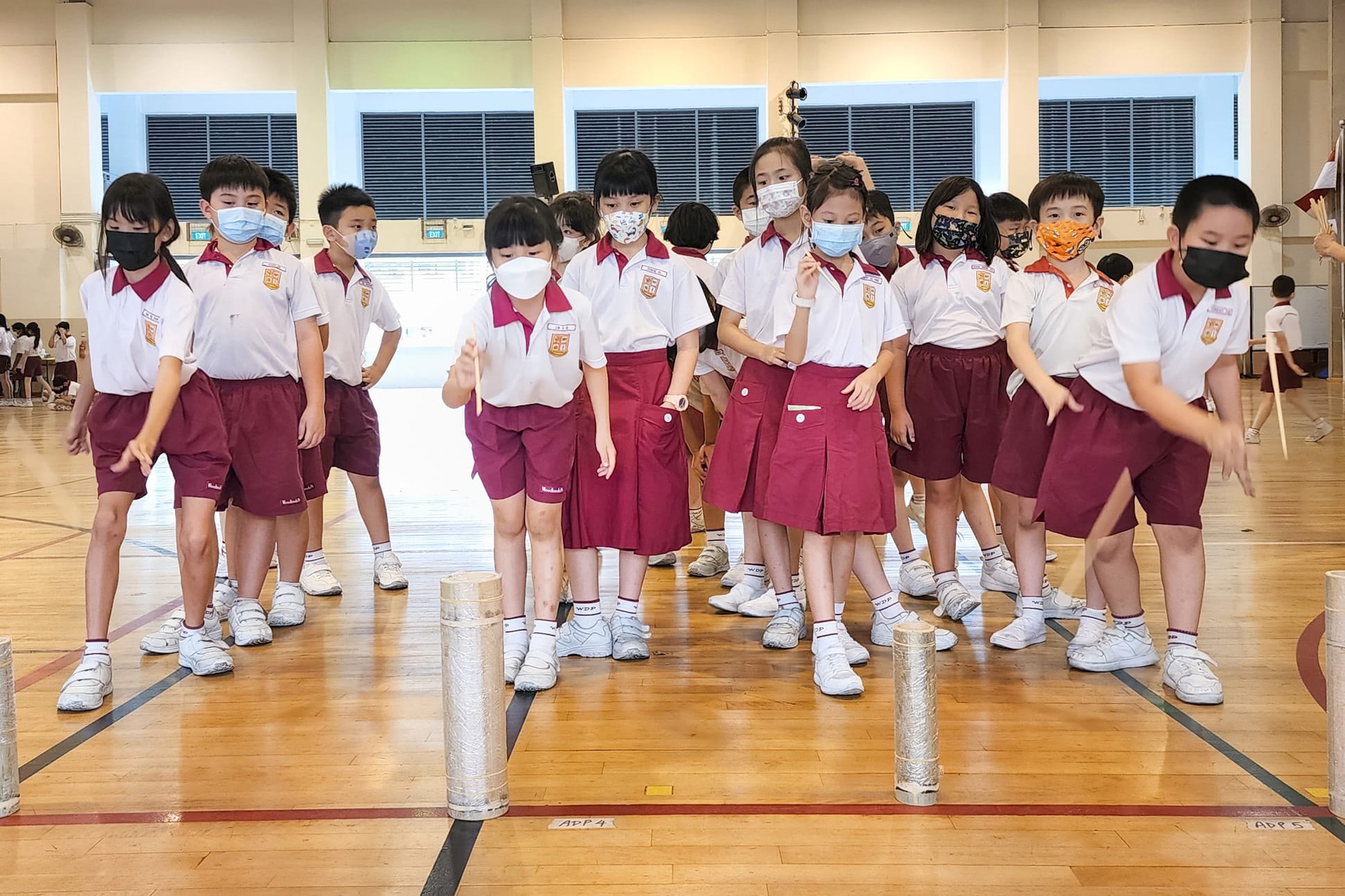 Young students in white and maroon uniforms wearing face masks stand in a school gymnasium near cylindrical objects.