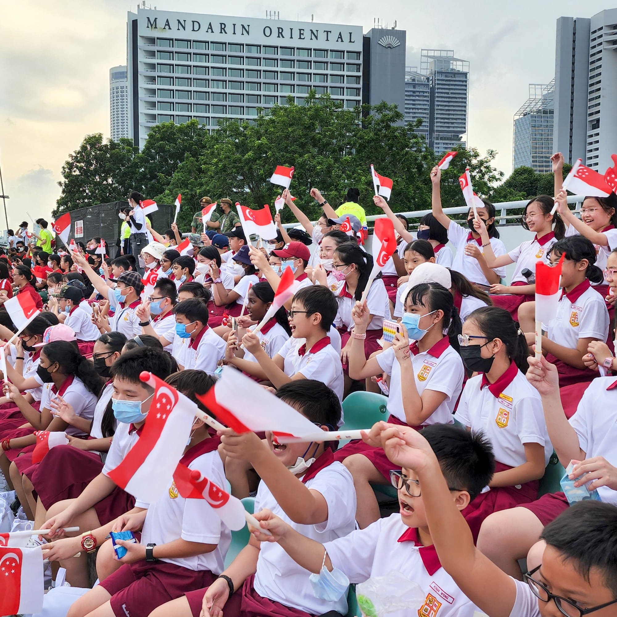 Students in white and red uniforms wave Singapore flags at an outdoor event near the Mandarin Oriental hotel.