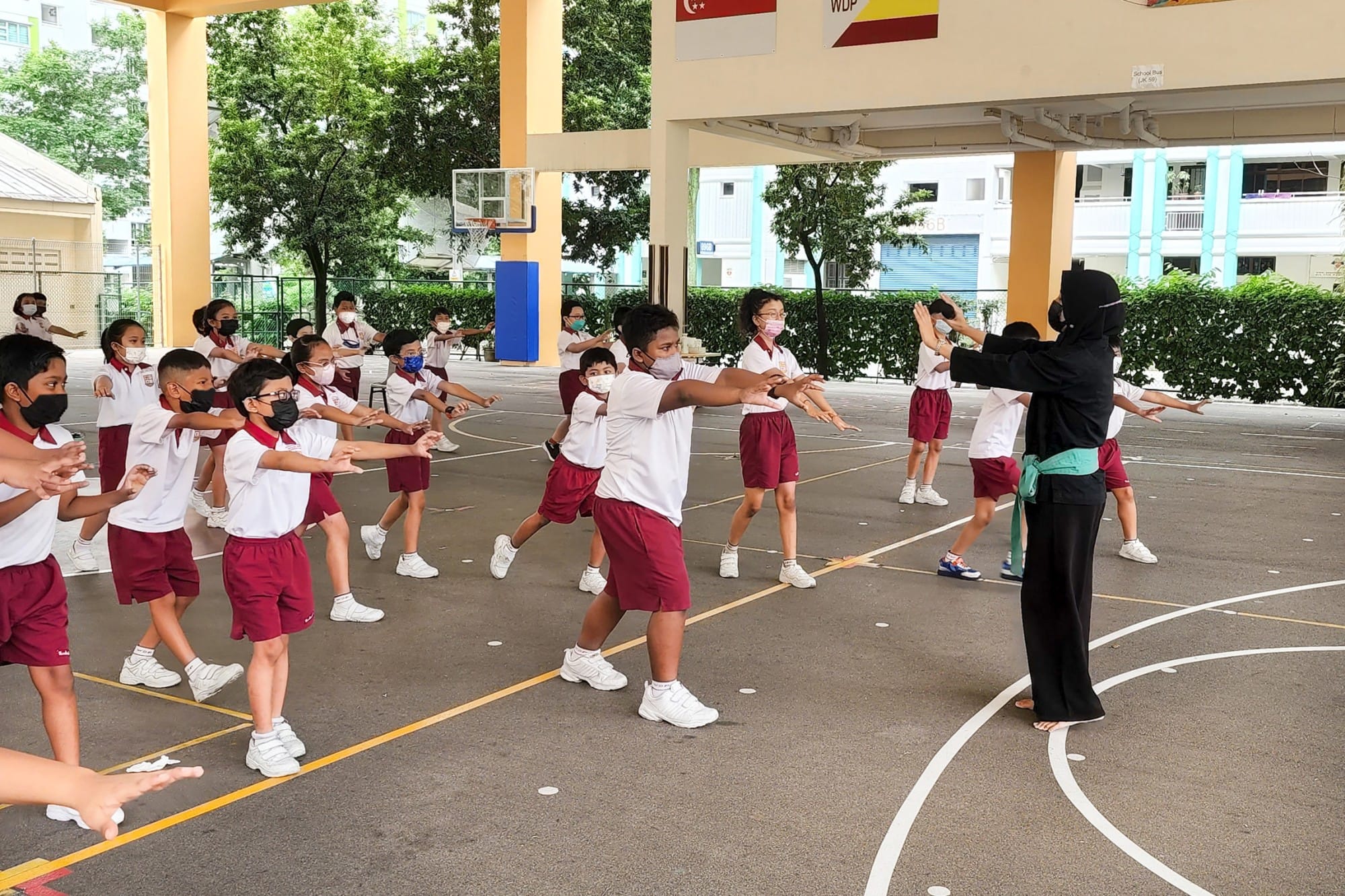 Children in school uniforms and face masks follow a teacher's exercise moves on an outdoor school court.